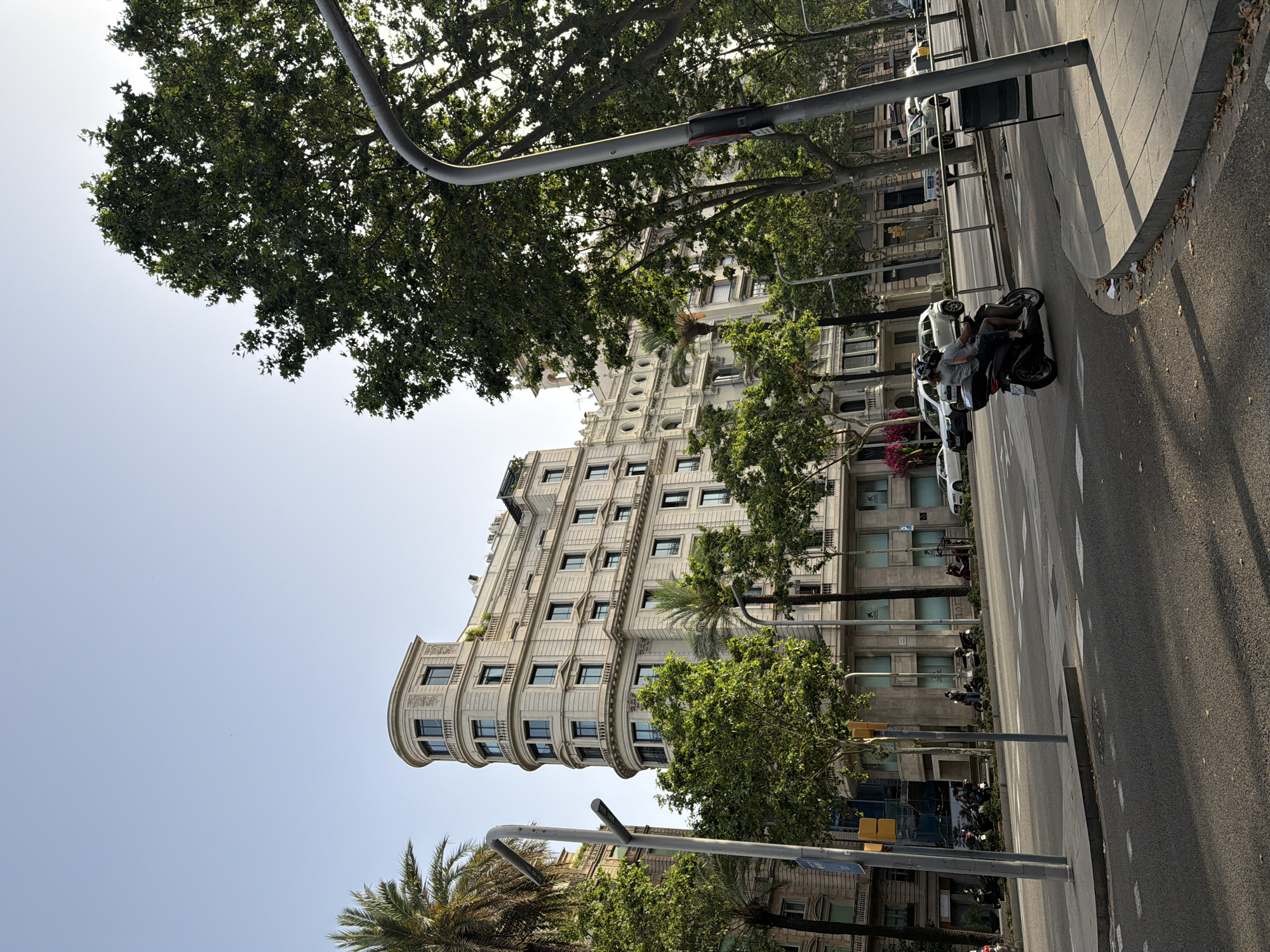 This is a street-level, eye-level, medium shot of a sunny day in Barcelona, Spain. The scene captures a curved building with light-colored stone facade and numerous windows on the left side, partially obscured by trees. In the foreground, a motorcycle with two occupants, wearing helmets, travels on the road. There are other vehicles and parked motorcycles visible further down the street, as well as trees lining the sidewalk and buildings in the background. The sky is clear and blue.