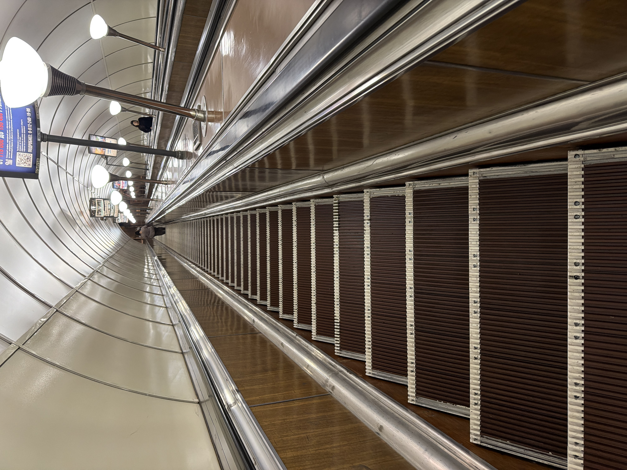 The image depicts a long escalator at the Avtovo metro station in Saint Petersburg, Russia. The escalator is viewed from above, looking down into the station. The walls are clad in light-colored panels with rows of spherical light fixtures extending along the left side. On the right, brown corrugated panels are visible along the length of the escalator, accompanied by a metal railing. The escalator itself has a brown wood-like surface with vertical white stripes. At the far end of the escalator, a few figures are visible. A sign with Russian text and a QR code is attached to one of the light fixtures. The scene appears to be indoors and well-lit.