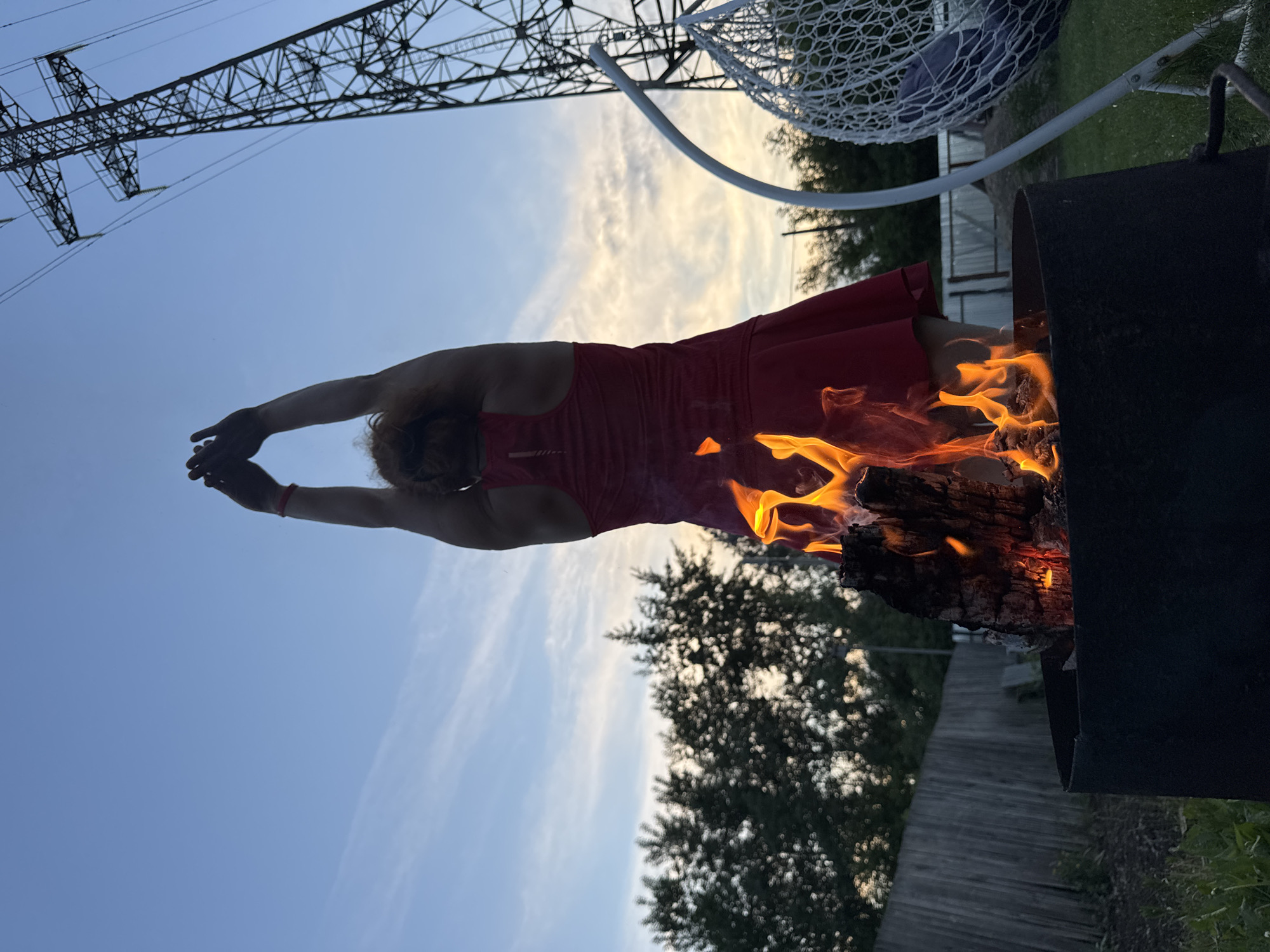 A person in a red dress stands in front of a burning log in a metal fire pit. The person's arms are raised above their head. In the background, there are trees, a wooden fence, a power line tower, and a white hanging chair. The sky is blue with some clouds. This scene is located in Chaikovsky, Russia.