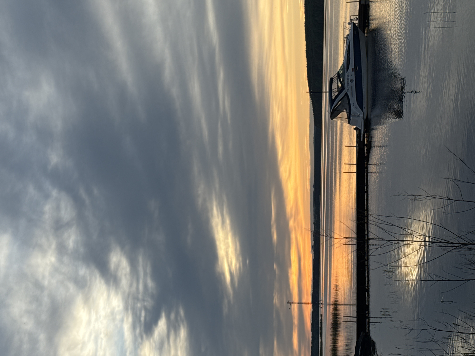 The image depicts a tranquil scene, likely taken at sunset, with a body of water reflecting the golden and orange hues of the sky. A boat is docked along a wooden pier. In the distance, a dark treeline is visible, contrasting with the bright sky. The presence of branches in the foreground suggests the photo was taken from a natural shoreline. The sky is filled with layered clouds, some dark and stormy, others catching the sunlight. I cannot confirm the location of this image within the city of Chaykovsky, Russia.