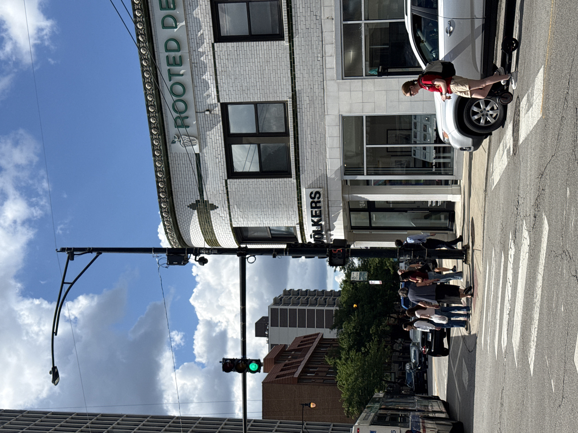 The image depicts a street scene in Chicago, Illinois, USA. A silver SUV is parked near a crosswalk where a young woman on an electric scooter stands. She wears a red shirt, khaki shorts, and glasses, with a backpack slung over her shoulder. The building behind her has "ROOTED DENTISTRY" signage and features white brickwork with decorative green trim. Across the street, a group of people waits at a bus stop, and a blue bus is visible. Traffic lights are illuminated green, indicating the flow of traffic. The environment is urban, with buildings of varying heights, trees, and cloudy skies.