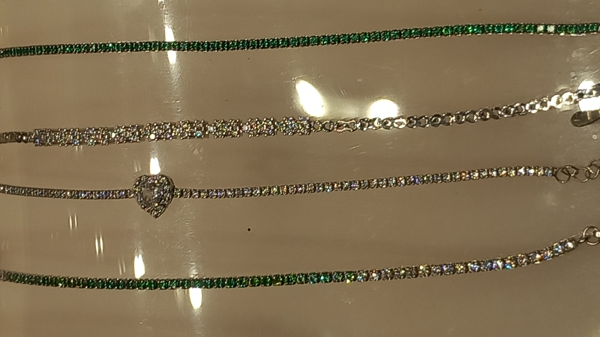 The image displays a close-up, vertical view of four bracelets arranged side-by-side, laid out on a light-colored, reflective surface, possibly glass or polished stone. The bracelets appear to be made of a silver-toned metal and are adorned with various stones.
From left to right:
1. The leftmost bracelet is a thin, delicate chain encrusted with small, green gemstones, likely emeralds or green cubic zirconia, giving it a vibrant appearance.
2. The second bracelet from the left is also a thin chain, but it features a larger, heart-shaped clear stone, possibly a diamond or clear cubic zirconia, centrally placed. The rest of the bracelet is adorned with small, clear, sparkling stones.
3. The third bracelet from the left is a moderately thick chain composed of interconnected links. It features decorative elements that resemble small, floral-shaped clusters of clear stones.
4. The rightmost bracelet is similar to the leftmost one, a thin chain filled with small, green gemstones, mirroring the first bracelet.
The background is a plain, light beige or cream color, with reflections of light sources visible as blurred oval shapes, suggesting overhead lighting. A small dark speck is visible on the surface between the first and second bracelets.
The image is an indoor product shot, likely in a jewelry store or display setting. There are no people or any indication of a specific outdoor location in the city of Kos, Greece. The overall impression is one of a clean, well-lit display of fine jewelry.