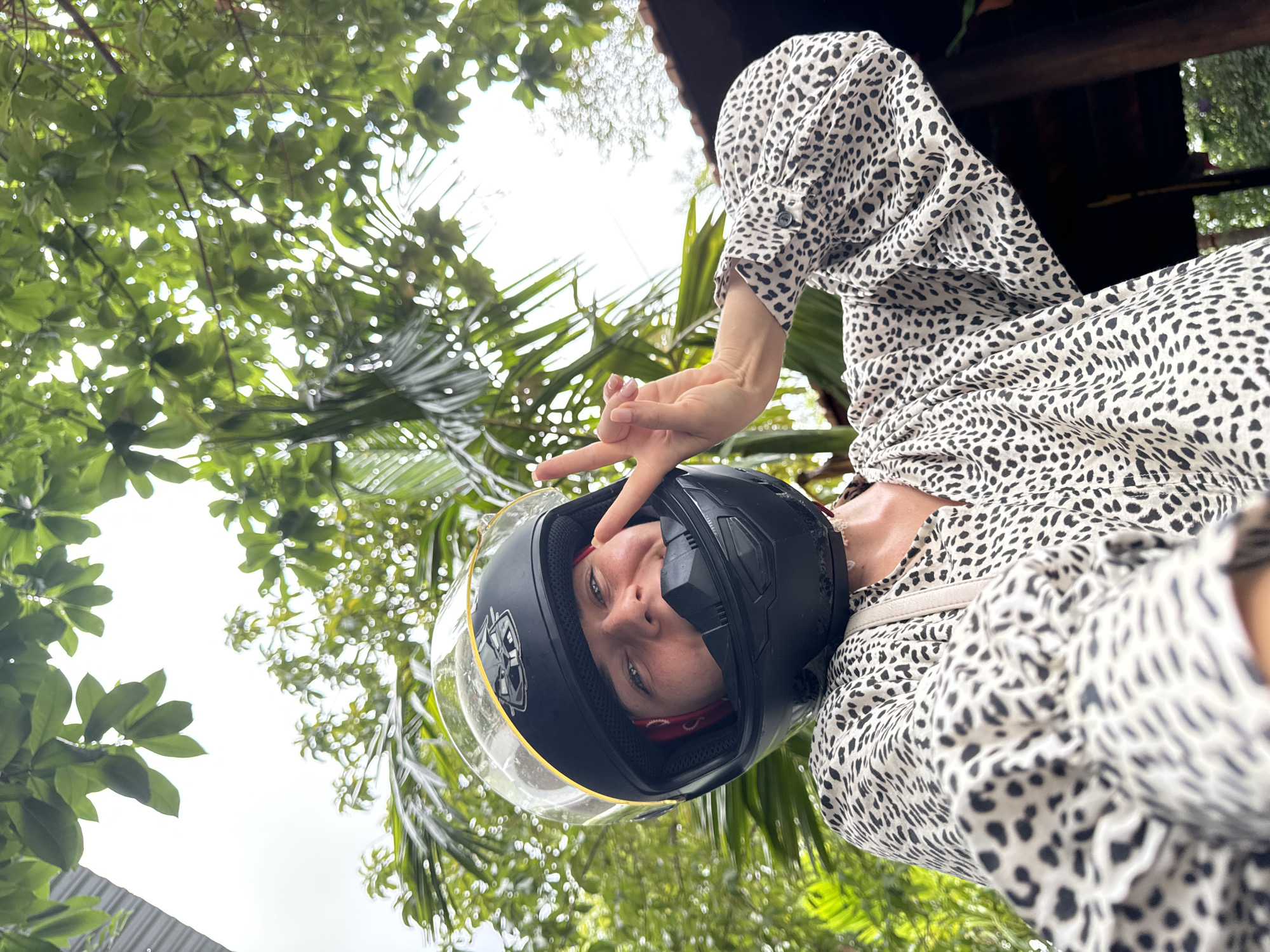 A person wearing a black motorcycle helmet with a clear visor and a tan leopard print dress is shown from the chest up, taking a selfie outdoors. The person is smiling and making a peace sign with their right hand. Lush green foliage, including palm leaves and larger tree leaves, frames the shot, with a bright, overcast sky visible through the leaves. A hint of a wooden structure is visible on the right side of the frame. The scene appears to be in Mũi Né, Vietnam, a coastal city known for its beaches and sand dunes.