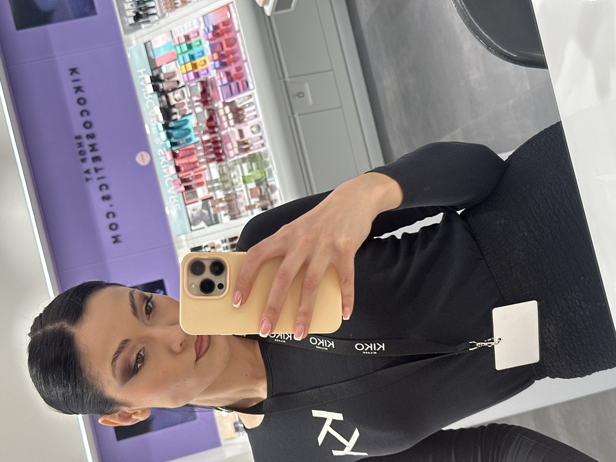 A woman is taking a selfie in a Kiko Milano store. She is wearing a black Kiko Milano shirt with a lanyard and a name tag. Her phone is a gold iPhone with a French manicure on her nails. Behind her, shelves are stocked with Kiko Milano cosmetics. The store is located in Burgas, Bulgaria.