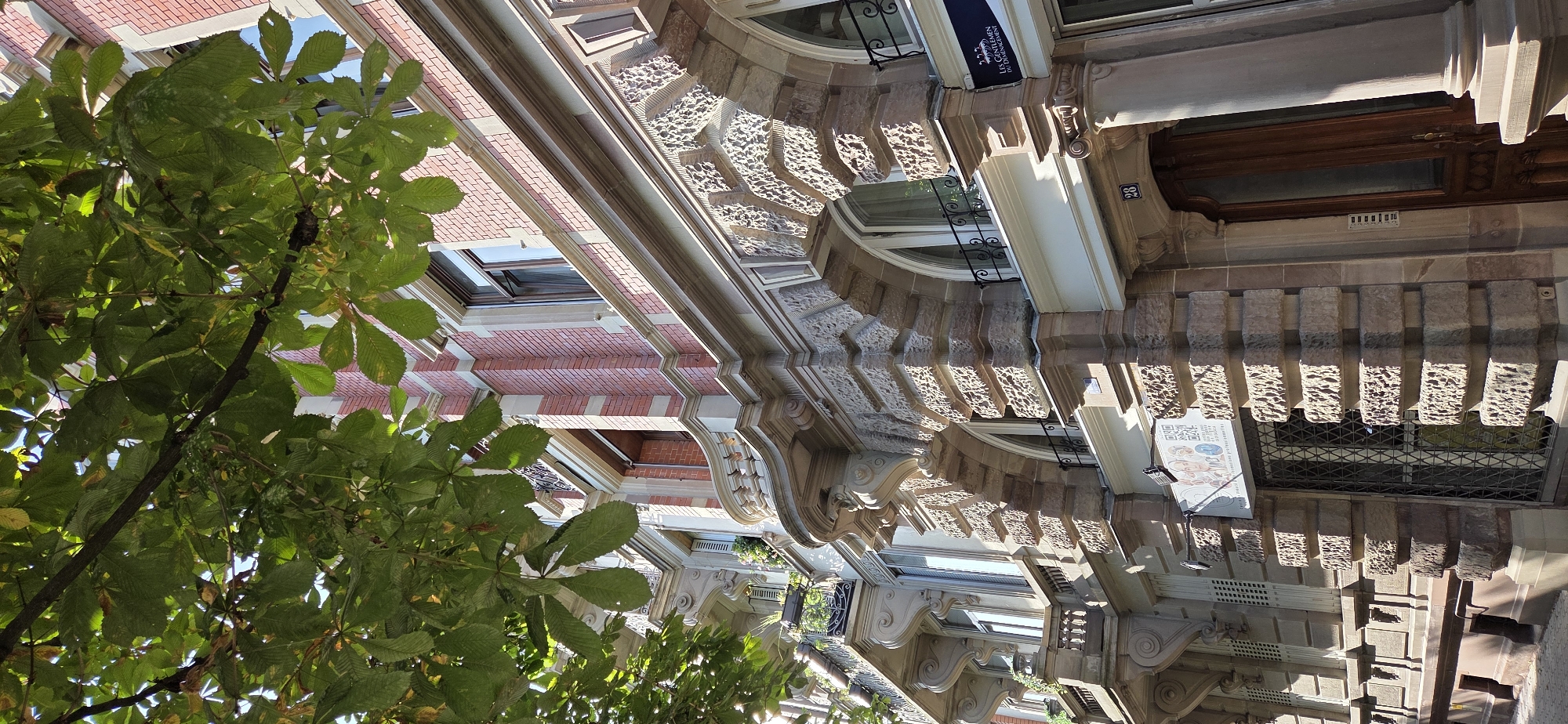 An exterior shot of a building facade is shown, with an overhead canopy of green leaves from a tree partially obscuring the upper portion of the image. The building is constructed with red brick on the upper floors and stone on the lower levels, featuring ornate architectural details. Windows with dark frames are visible, some with balconies. The stone detailing includes carved, rough-hewn blocks that form decorative arches and cornices. On the ground level, a large wooden door with glass panes is visible, next to a metal-grated window. A sign with French text, "Les Gentlemen du Déménagement," is mounted on the facade. The overall impression is of an older, well-maintained building in an urban setting.