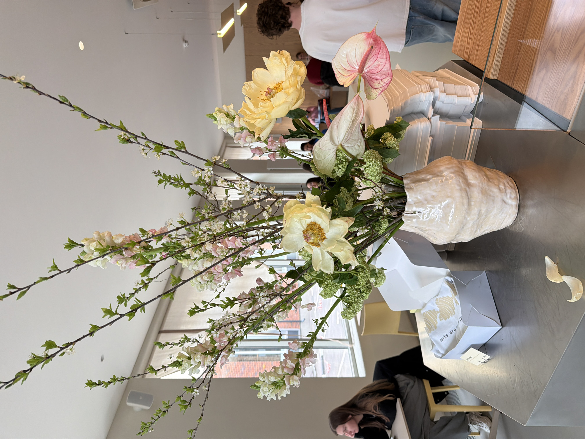 The image shows a floral arrangement in a ceramic vase, placed on a metallic table. The arrangement includes yellow peonies, white anthuriums, and branches with small white and pink blossoms. Near the vase is an open white box with printed text "carbs are." The background includes a partially visible window with a brick building visible outside, as well as glimpses of people in the interior space. Moscow, Russia is not evident in the photograph.