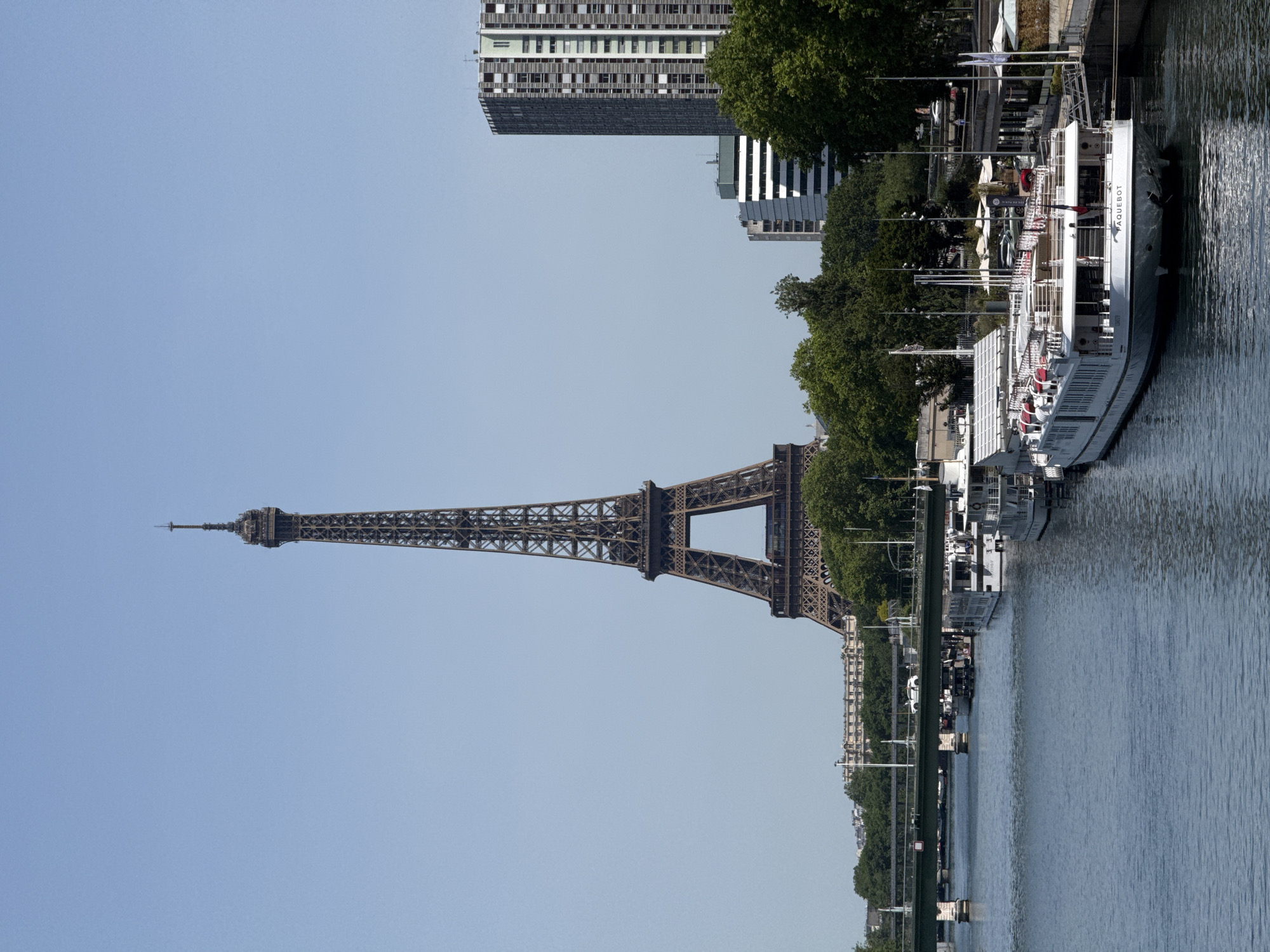 The image features the Eiffel Tower in Paris, France, seen from a lower vantage point. The tower is prominently displayed against a clear blue sky. Along the right side of the image is the Seine River, where several boats are docked. A building is seen in the background.