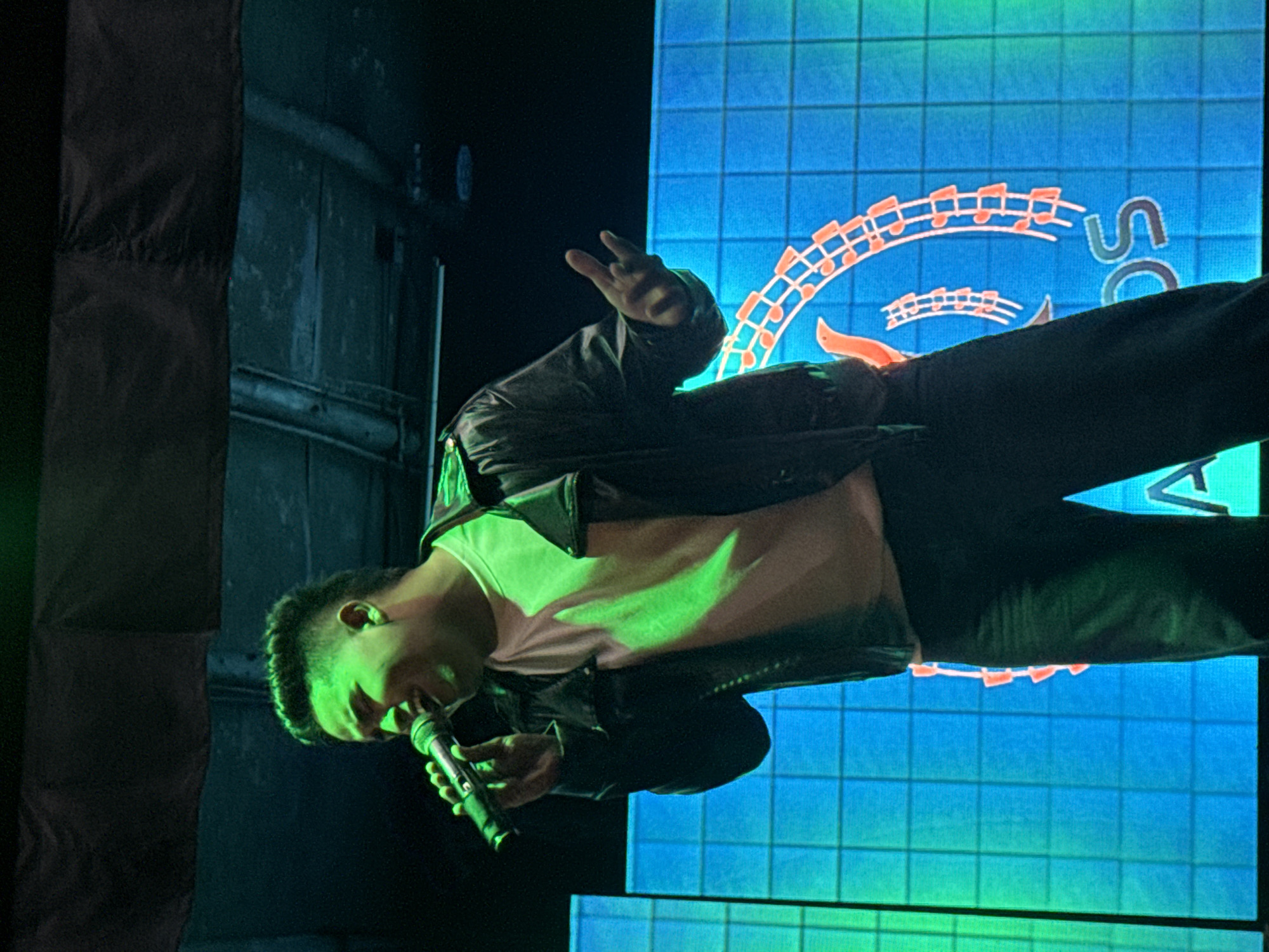 A man is singing into a microphone on a stage in front of a blue screen with orange musical notes and the letters "SC." He is wearing a black jacket and pants, and a white t-shirt. He is holding his left hand in a "peace" sign. The environment is dark with a spotlight shining on the performer. There is a black object to the left of the performer, possible some sort of equipment. There are no indications of the city of Porlamar, Venezuela.