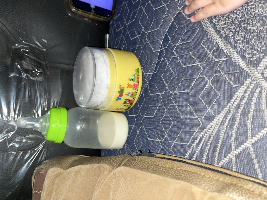The scene depicts baby-related items on a patterned surface, located in Tempel, Indonesia. A clear baby bottle with a bright green screw-on cap and nipple contains approximately one-third full of white liquid, consistent with milk or formula. Adjacent to the bottle is a yellow cylindrical container with a clear plastic lid, identified by the label "Yuki" and a colorful cartoon graphic of a train carrying animals. The container appears to hold a white powdery substance, visible through the lid. Both objects rest on a blue fabric surface with a repeating light-colored geometric pattern resembling interconnected hexagons.
To the left of the baby bottle, a portion of a light brown upholstered object, possibly a sofa armrest or cushion, is visible, featuring a visible zipper and textured material. In the background, behind the bottle and container, is a dark, possibly black, surface, partially covered by crumpled clear plastic sheeting. On the far right, a sliver of a smartphone or tablet screen, displaying a blue image, is discernible within a clear case. In the bottom right corner of the frame, the fingers and partial palm of a small, light-skinned hand, consistent with that of a child, are resting on the patterned blue fabric. The overall lighting is dim.