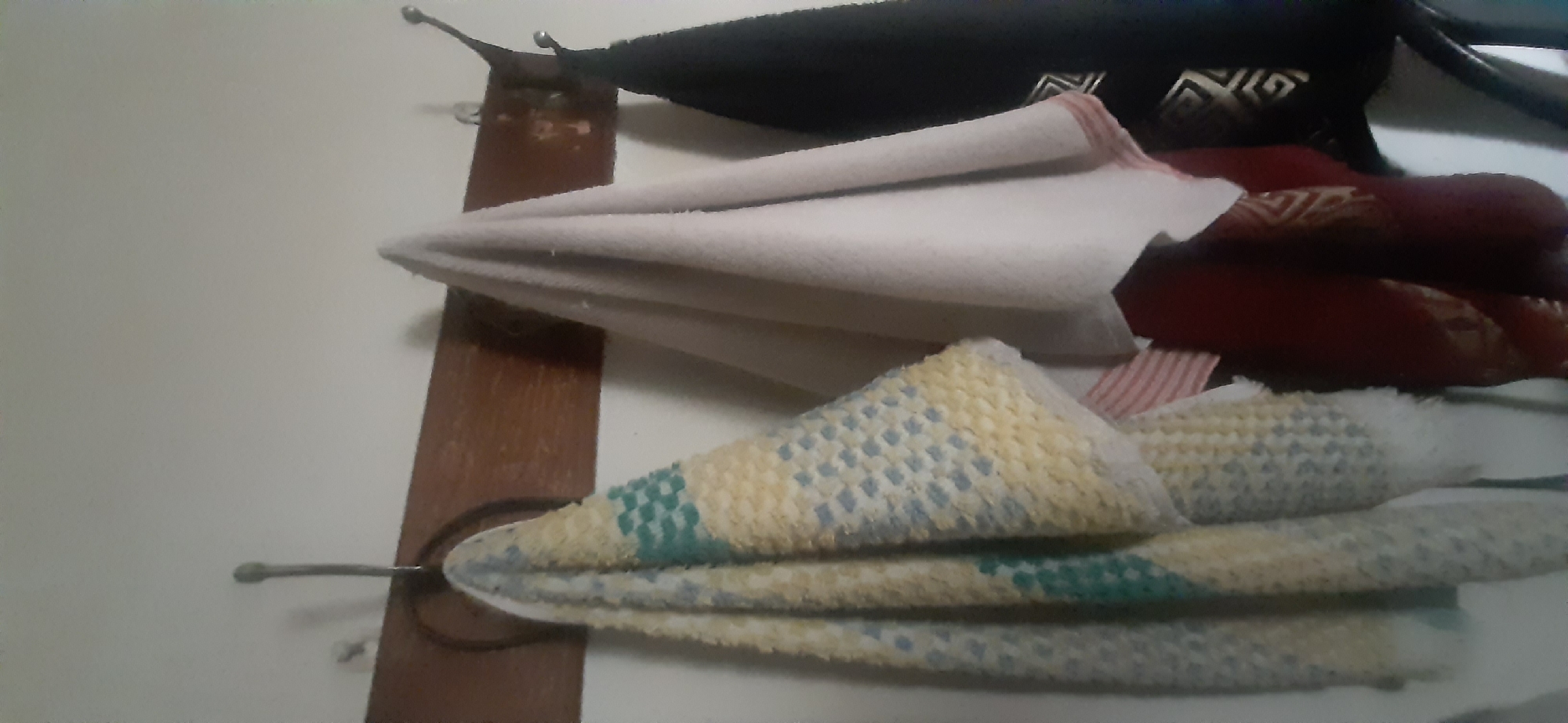 The image shows a wooden rack mounted on a light-colored wall, with three towels hanging on it. The leftmost towel is yellow and blue, with a checkered pattern. The middle towel is a light cream color with red stripes along the bottom edge. The rightmost towel is black with a gold geometric pattern. The towels appear to be in a domestic setting, possibly a bathroom or kitchen. There is no information to suggest this image is related to Yessentukskaya, Russia.