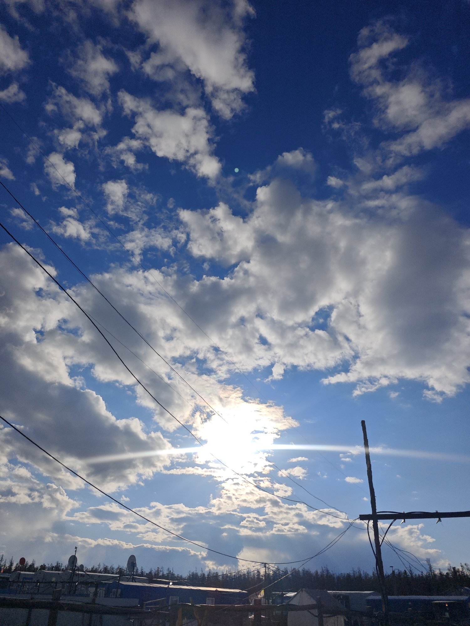 The image depicts a daytime scene in what appears to be a residential area in Unknown, Russia, characterized by a bright, cloudy sky. The sky is predominantly blue, interspersed with numerous white cumulus clouds. The sun is visible, partially obscured by the clouds, creating a bright glare.
Several black power lines run diagonally across the frame, intersecting with the sky and clouds. Below the horizon, a cluster of buildings and structures are visible, including satellite dishes on the roofs. A vertical wooden pole with attached wires stands out against the sky. A dense row of trees lines the horizon behind the buildings. The overall scene conveys an ordinary day in a populated area with typical infrastructure.