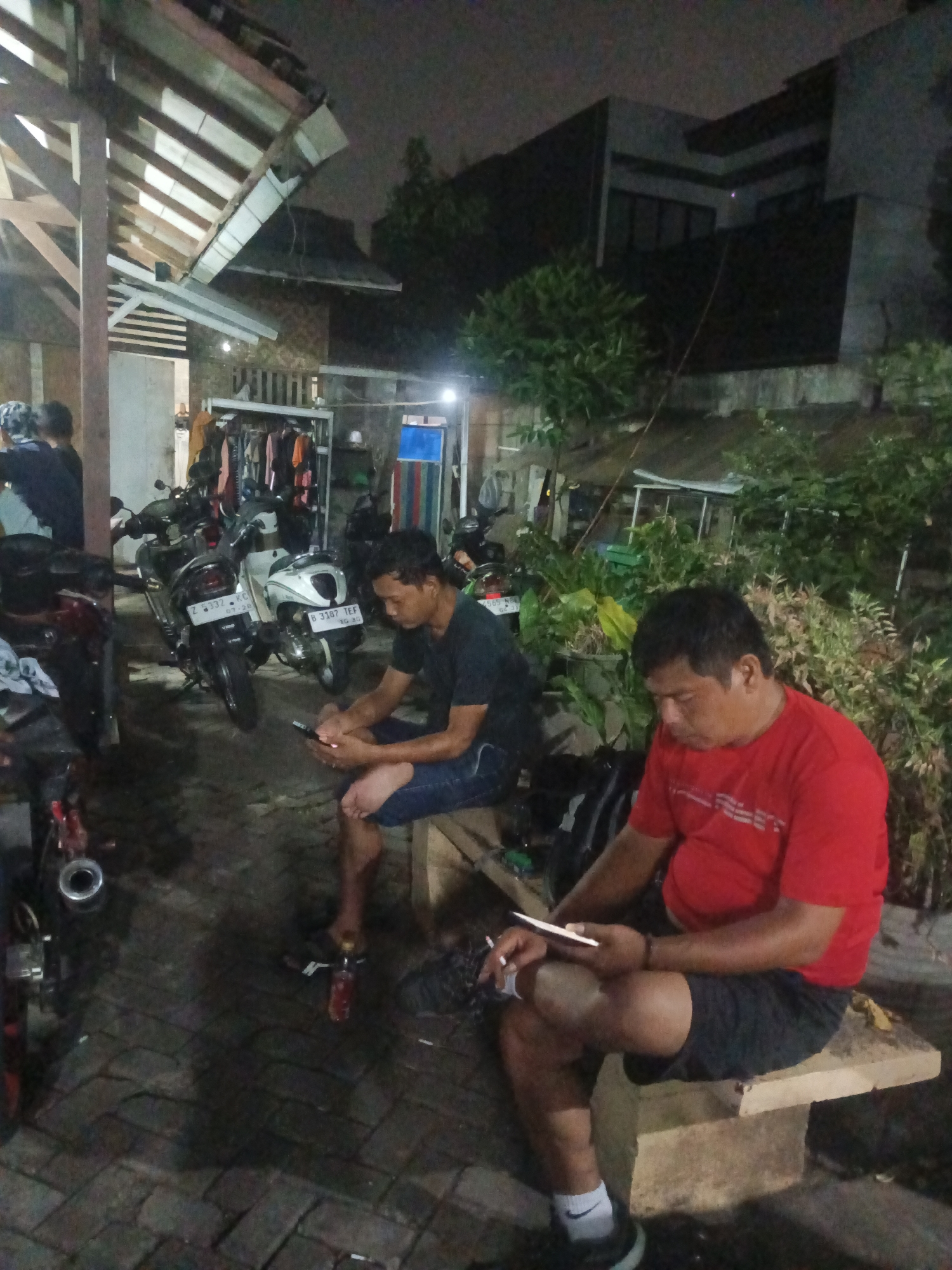 The image shows a dimly lit outdoor scene, likely in the city of South Tangerang, Indonesia, at night. In the foreground, two men are seated. The man on the left, wearing a dark t-shirt and blue jeans, is looking at his mobile phone. To his right, another man in a red t-shirt and dark shorts is also looking at his phone, with a cigarette in his left hand. Both men appear to be sitting on makeshift seating.
Behind them, several motorcycles are parked, suggesting a public space or a gathering area. In the background, there is a structure that appears to be a building with a tiled roof and some exposed wooden beams. Lush green foliage surrounds the area, indicating a tropical environment. The overall atmosphere is casual and relaxed, with the men engaged with their devices. The ground is paved with stone tiles. A bottle of what appears to be a beverage is visible near the feet of the man on the left.