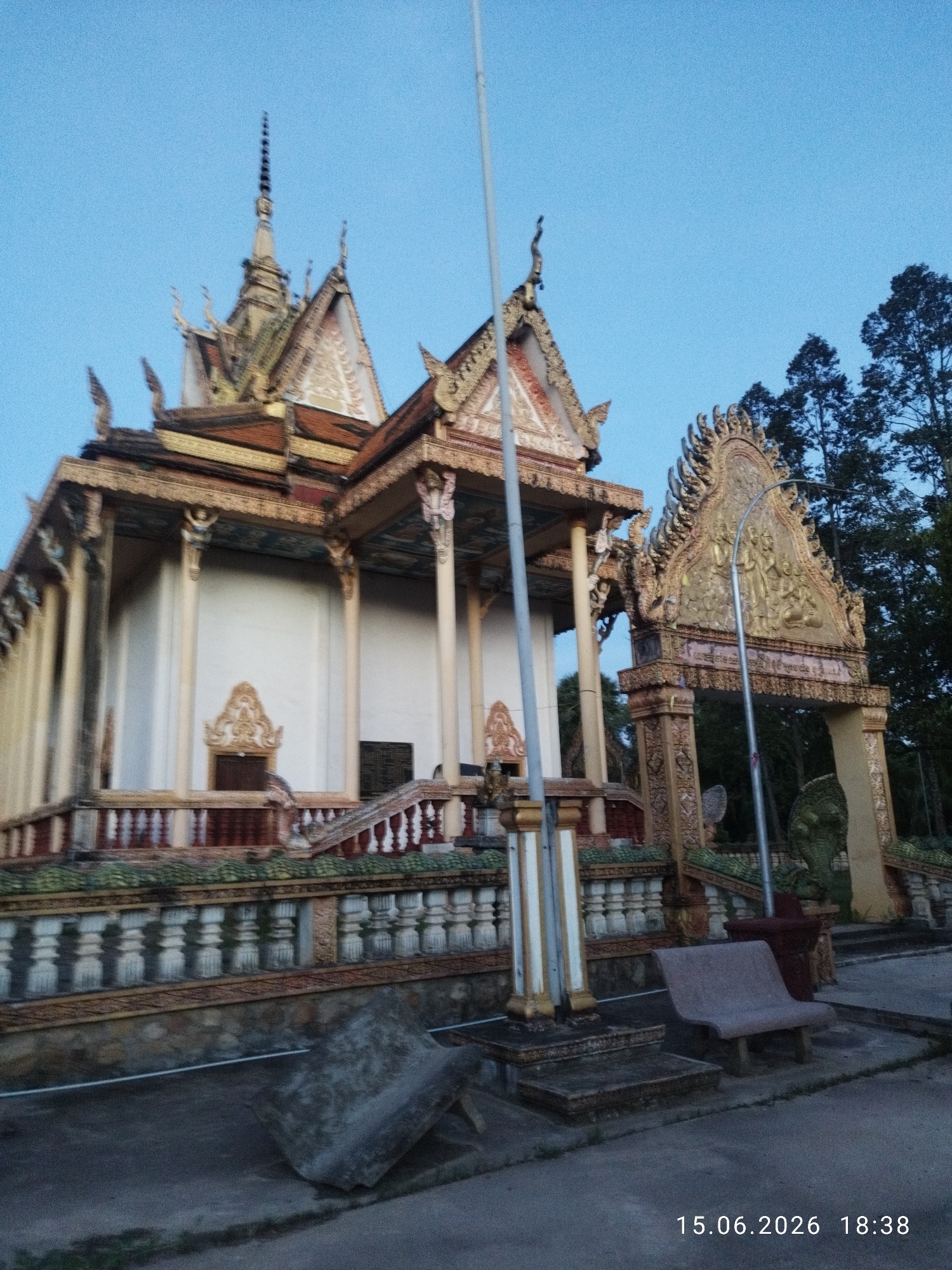 The image depicts a traditional Cambodian temple or pagoda located in Kampot, Cambodia. The structure is adorned with intricate golden carvings and ornate roofs, characteristic of Khmer architecture. Pillars support the roofed entrance, and a multi-tiered railing with decorative elements lines the front of the building. Behind the main structure, tall trees are visible against a clear blue sky. In the foreground, there's a concrete bench and what appears to be a large, rough-hewn stone slab. A streetlamp stands to the right of the entrance archway, which is also elaborately decorated with golden reliefs. The scene is captured in daylight, with the timestamp "15.06.2026 18:38" visible in the lower right corner. The overall impression is one of a serene and culturally significant site.