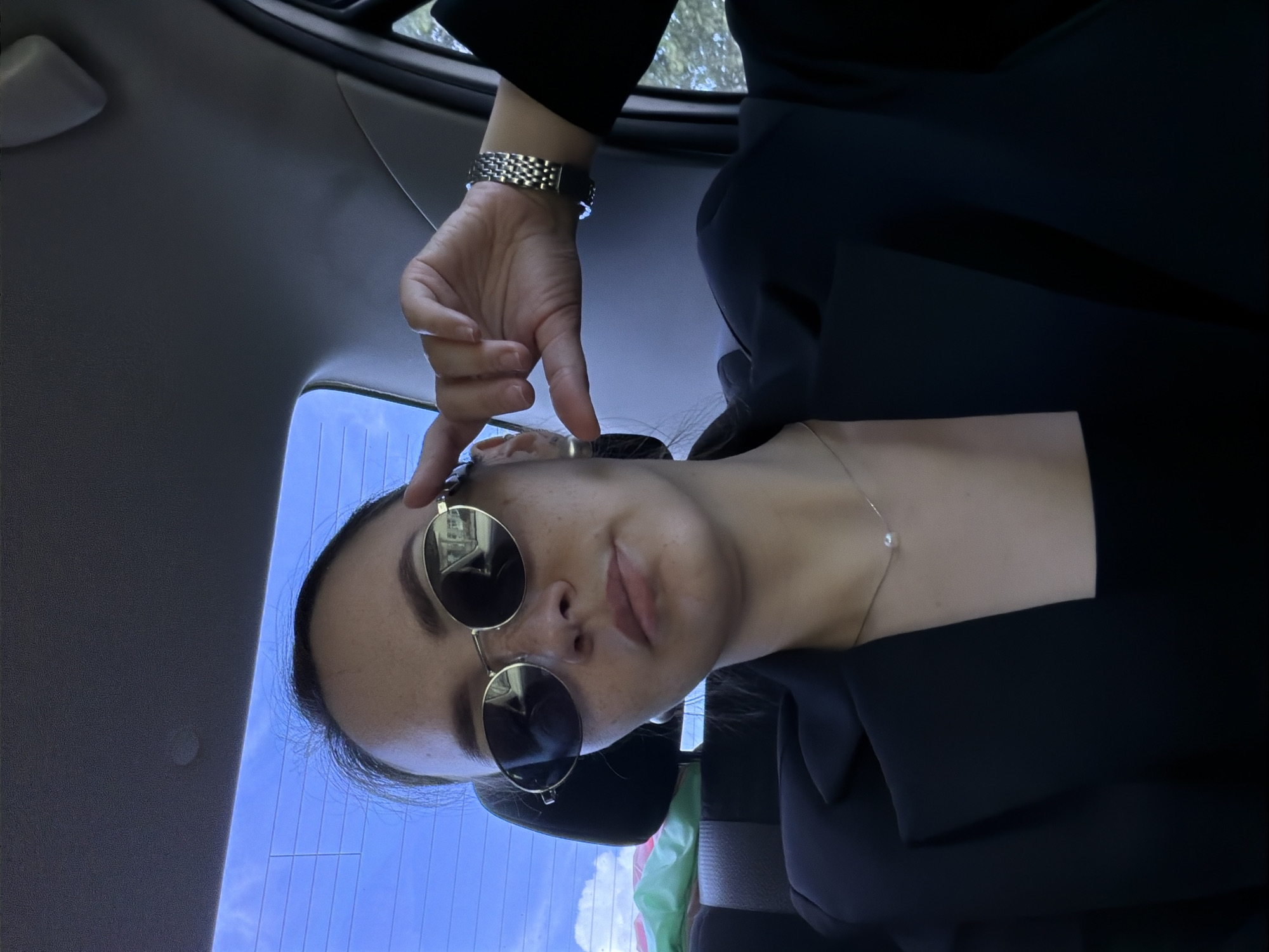 A young woman with dark hair pulled back, wearing sunglasses, a black blazer over a black top, and a pearl necklace, is photographed from the chest up inside a car. She is looking at the camera and adjusting her sunglasses with her right hand. The background shows the rear window of the car with a blue sky and some clouds visible outside. There is a headrest of a car seat and some green and pink fabric objects in the back seat. The woman is likely in the city of Chaikovsky, Russia, as indicated by the context.