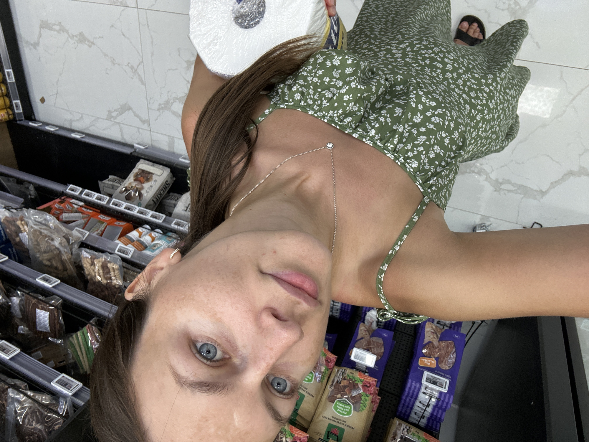 A young woman with blue eyes and brown hair, wearing a green floral print dress and black sandals, is taking a selfie in a grocery store in Donji Pobori, Montenegro. She is holding a roll of toilet paper. The background shows shelves stocked with various products, including pet food. The floor is tiled with a marble-like pattern. The lighting suggests an indoor environment with artificial lights.