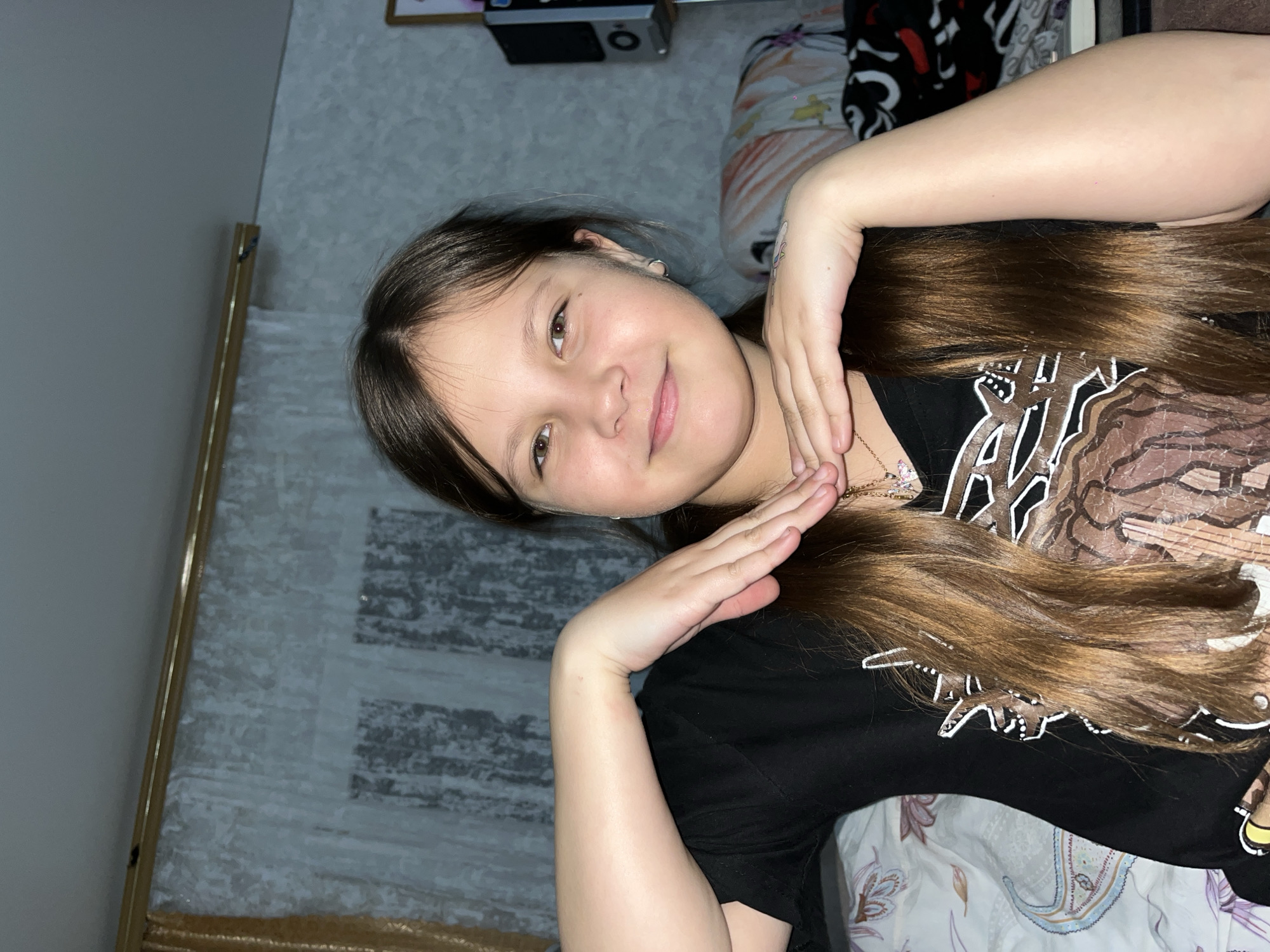 A young Caucasian girl is lying down, looking at the camera. Her brown hair is spread out around her. She is wearing a black t-shirt with a white and brown design. Her hands are raised to her neck and face in a posing manner. The background includes a patterned wall, part of a bed with patterned sheets, and an object in the upper portion of the image that appears to be a device of some sort. There is no clear information to indicate the image was taken in Novopokrovskaya, Russia.