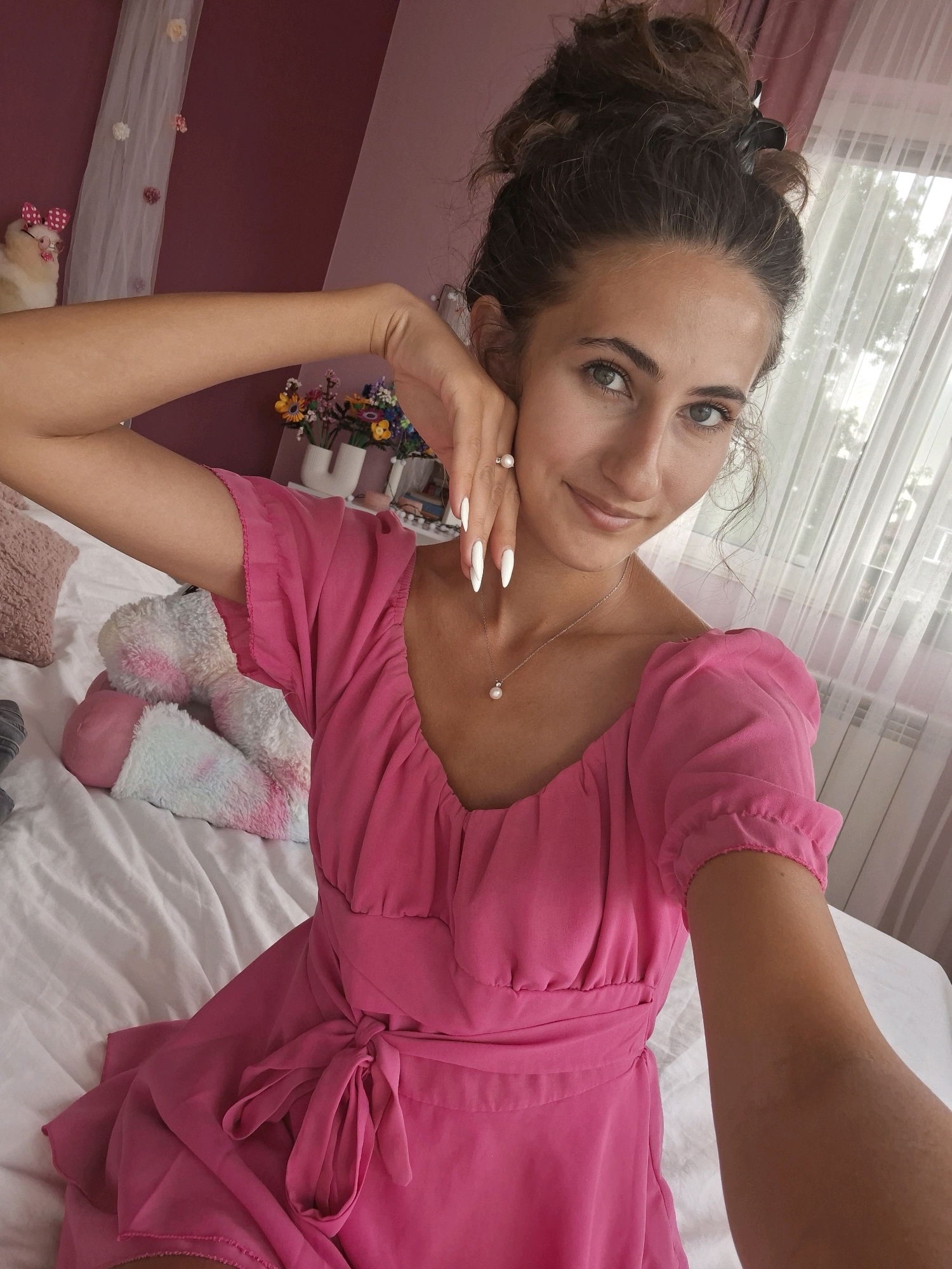 A young woman with dark, curly hair styled in a messy bun is taking a selfie in a bedroom. She is wearing a bright pink dress with puffed sleeves and a tie-front detail. Her left arm is raised, with her hand resting on her cheek. She has long, white painted nails and is wearing a pearl necklace and a ring with a pearl. The background shows a white-covered bed, a fluffy, pastel-colored stuffed animal, a shelf with small potted flowers, and a sheer white curtain covering a window. The wall is painted a muted pink.