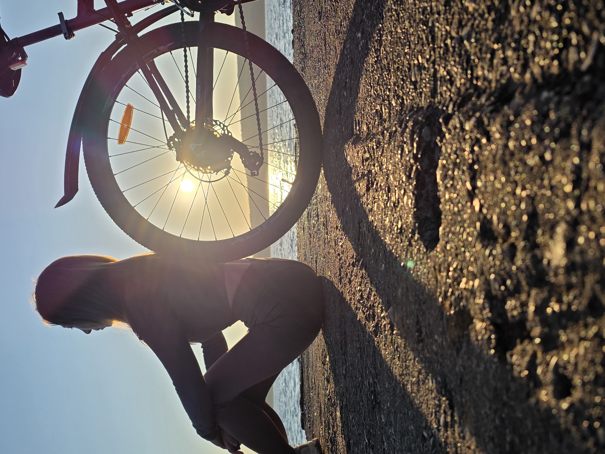 A silhouette of a woman sitting next to a bicycle on a paved path by the water is captured during sunset. The sun is visible through the spokes of the bicycle's rear wheel, casting a warm glow on the scene. The woman is wearing athletic clothing and is facing away from the camera, looking out at the water. The path is made of gravel and asphalt, and there are tire tracks visible in the foreground. The water is calm, and the sky is clear blue with a hint of orange from the setting sun. The location is the embankment of the Kama River in Chaikovsky, Russia.
