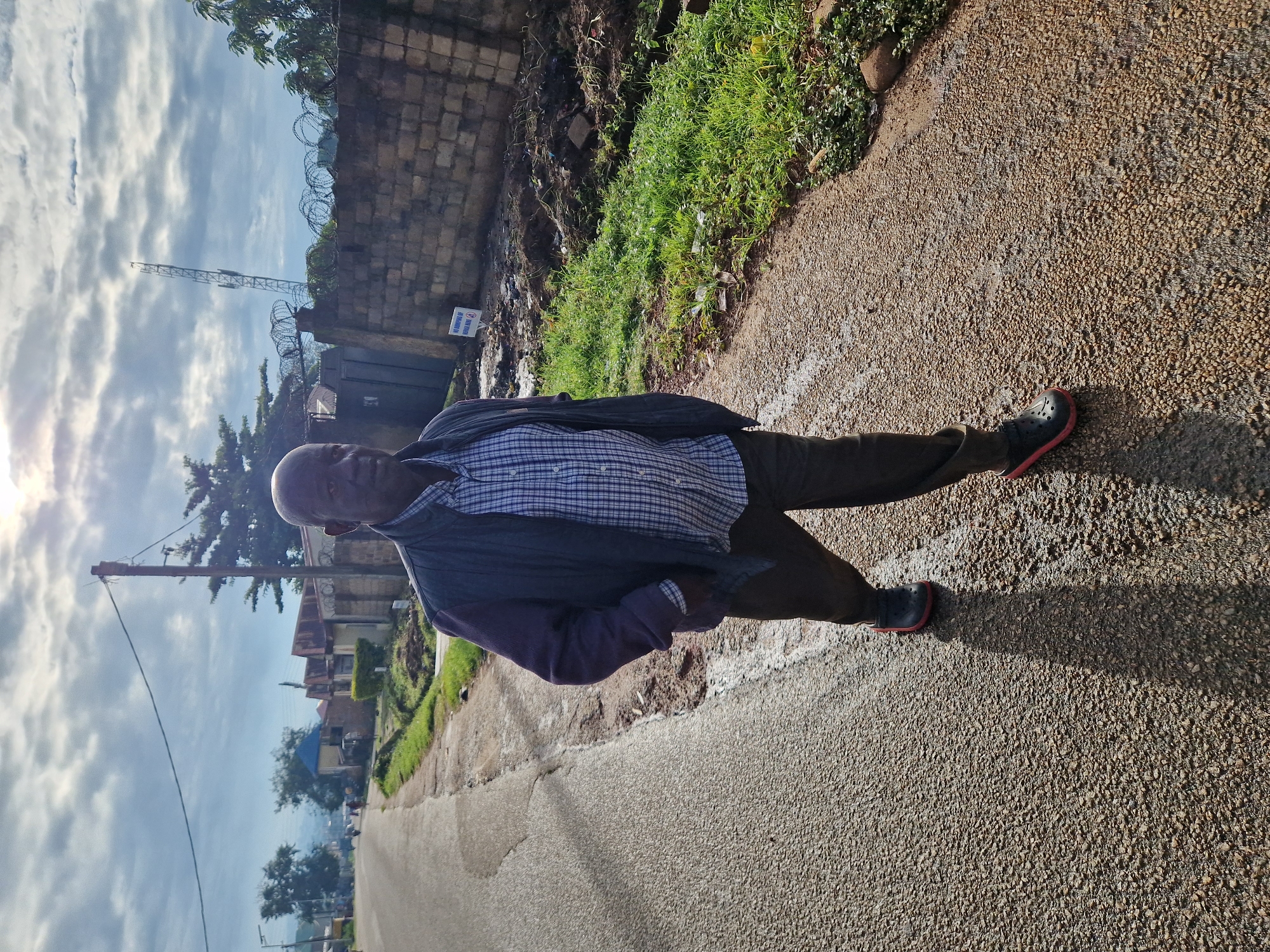 A man is walking on a wet asphalt road with gravel on the side, in the city of New Stadium, Nigeria. The man is wearing a blue and white checkered shirt, a dark jacket, dark pants, and black and red crocs. He is walking towards the camera, with his right hand in his pocket. The road is lined with green grass and a brick wall on the right side of the image. Behind the wall, there are some buildings and a telecommunications tower. The sky is cloudy with some sun peeking through. There is a sign on the wall that says "NO DUMPING OF REFUSE HERE".