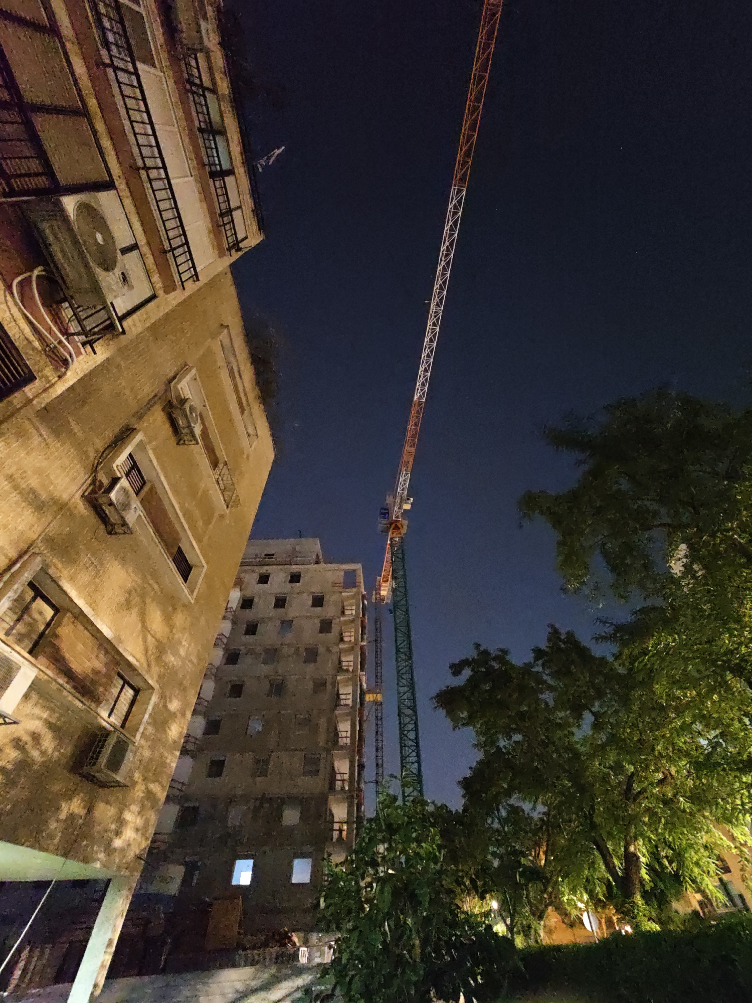 The photograph, taken at night in Petah Tikva, Israel, captures a construction site adjacent to residential buildings. The scene is dominated by a tall construction crane, its lattice structure visible against the dark blue night sky. The crane's boom extends diagonally upwards, segmented by orange and white paint. To the left of the crane, a multi-story building with a worn, textured facade is partially visible. This building features several windows, some of which are dark, while others have internal light sources, suggesting occupancy. Air conditioning units are mounted on the exterior walls, and metal fire escapes run vertically alongside the windows. Further into the background, another tall, unfinished building is visible, its numerous windows appearing as dark openings. Green trees with dense foliage are positioned in the foreground and to the right of the crane, illuminated by a warm light source, likely from streetlights or the construction site itself. The overall impression is one of urban development occurring in close proximity to established residential areas, under the cover of darkness.
