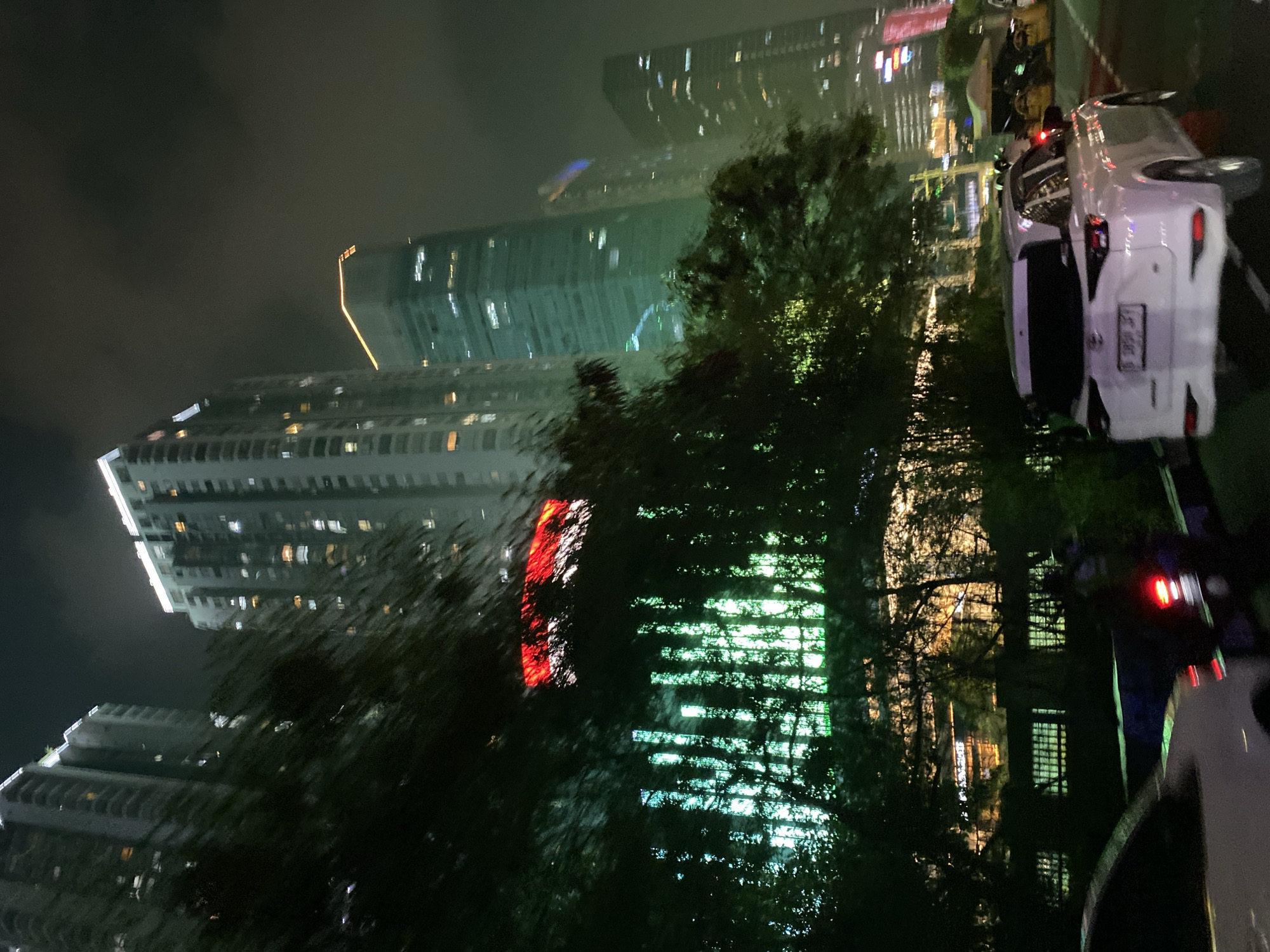 The image shows a nighttime street view in Tangerang, Indonesia, with several tall buildings in the background. A white SUV is parked in the foreground, facing away from the camera. Trees and foliage are visible on the left side of the image, partially obscuring some of the buildings. The buildings are illuminated with lights, some of which display red and green patterns. The overall atmosphere is urban and modern, with a sense of activity due to the visible lights and vehicles.