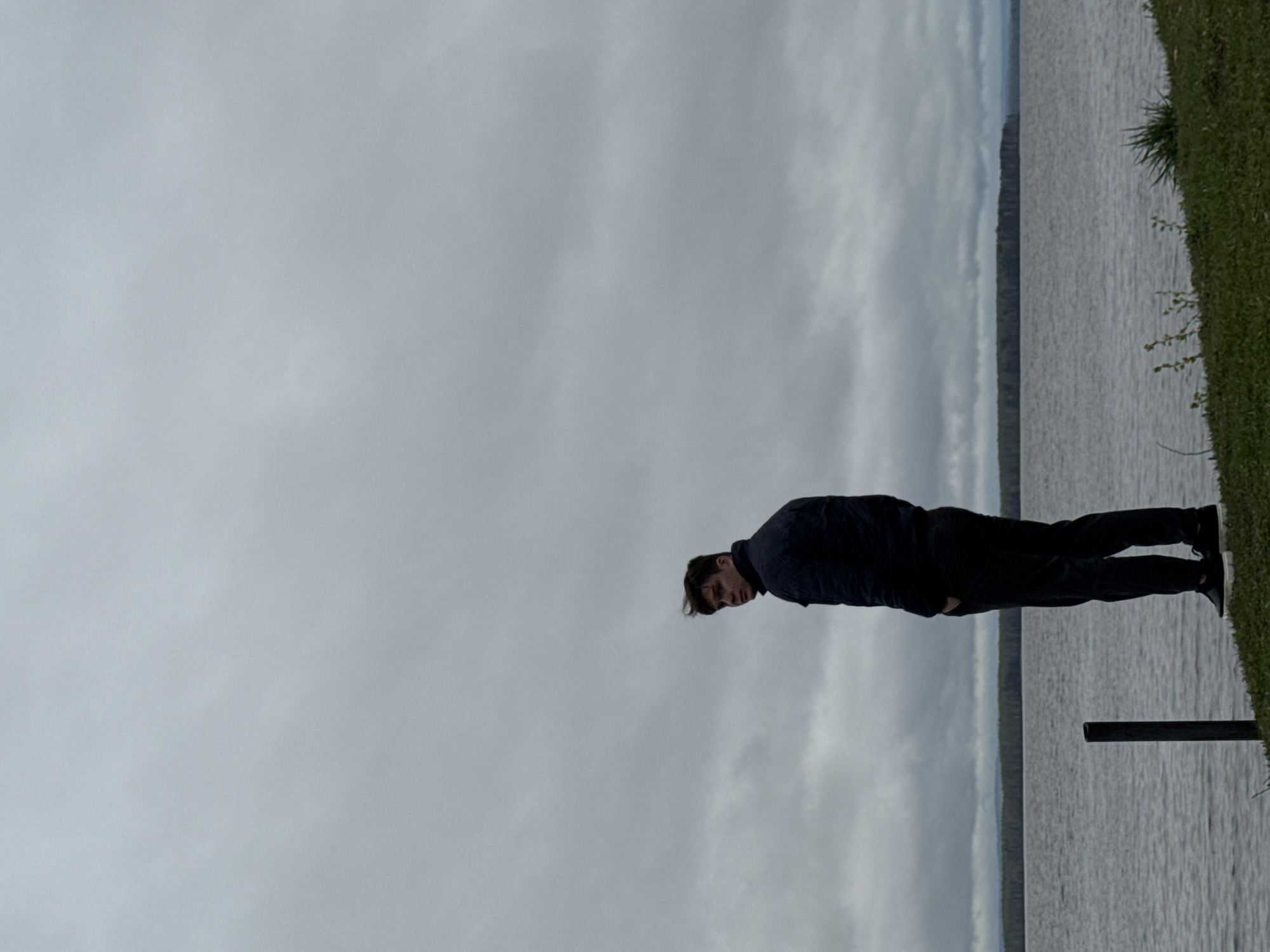The image shows a man standing near a body of water, likely the Kama River in Chastyye, Russia. The man is wearing dark clothing, a jacket and pants, and black shoes with white soles. He is positioned on a grassy area near the water's edge. A metal railing is visible behind him. The sky is overcast, with grey clouds. The water appears calm, with a faint line of trees visible on the opposite shore.