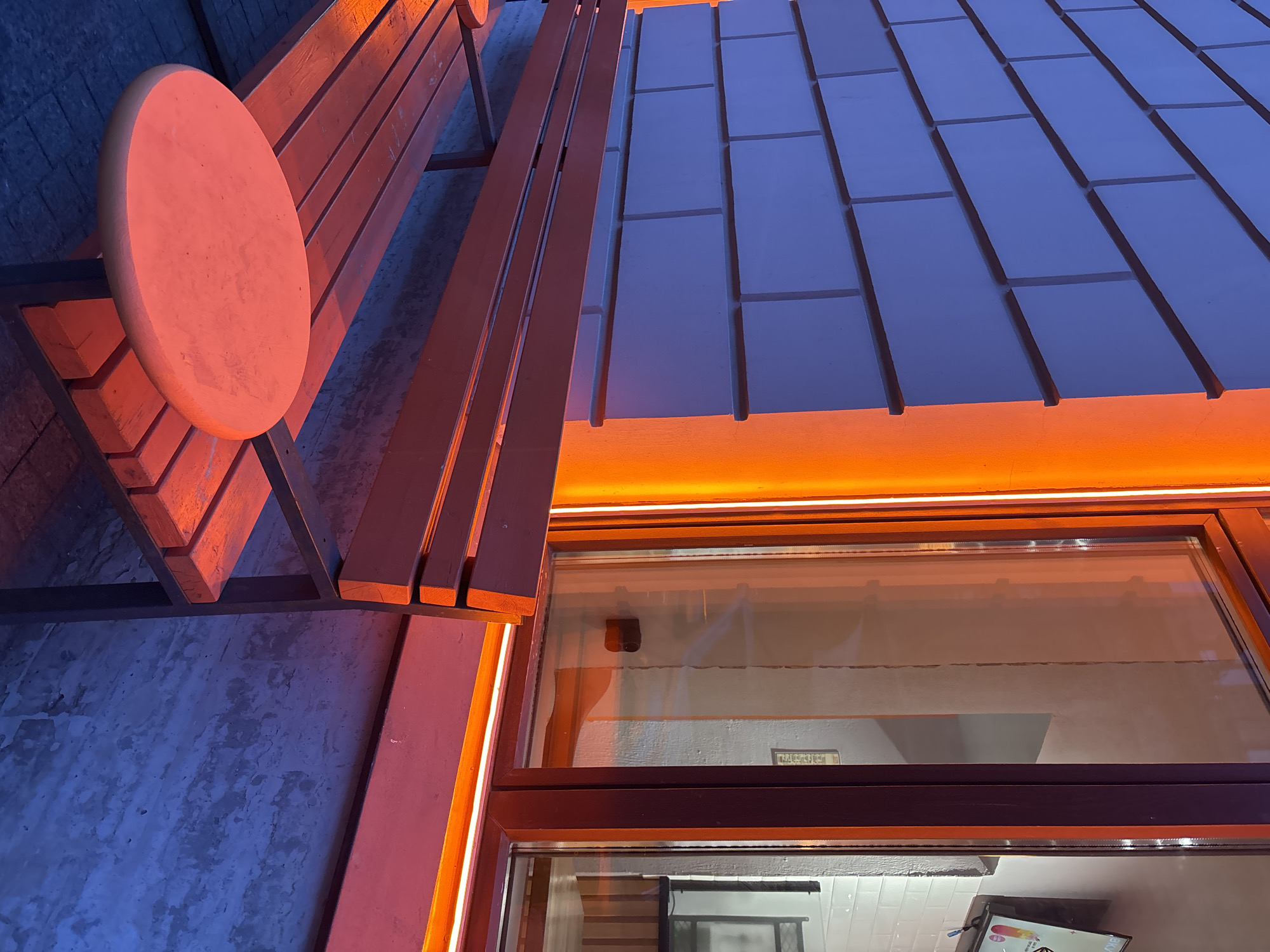 This image shows a brightly lit street scene in Saint Petersburg, Russia, featuring a modern building facade with a long, rectangular awning composed of dark gray, slightly glossy panels. The awning is illuminated from below with warm, orange light, which is also reflected in a large window beneath. The window's dark brown frame matches the wood used in a nearby bench with a circular table attached to one end. The bench is positioned next to the building and is made of light-colored wood. The ground surface is a mottled gray, possibly concrete or stone. The overall atmosphere is warm and inviting, likely photographed at dusk or at night.