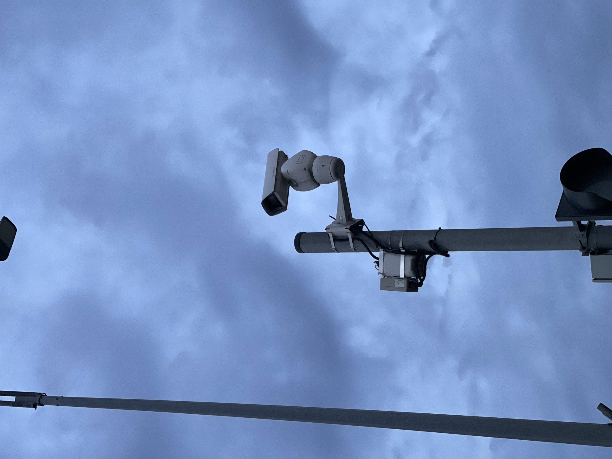 A security camera is mounted on a pole, pointed forward and slightly upward against a cloudy sky. A second, smaller box is mounted below the camera. A tall, thin pole stands to the left of the camera pole, reaching from the bottom to the top of the frame. A traffic signal is visible at the bottom right corner of the image. The overall color palette is muted blues and grays.