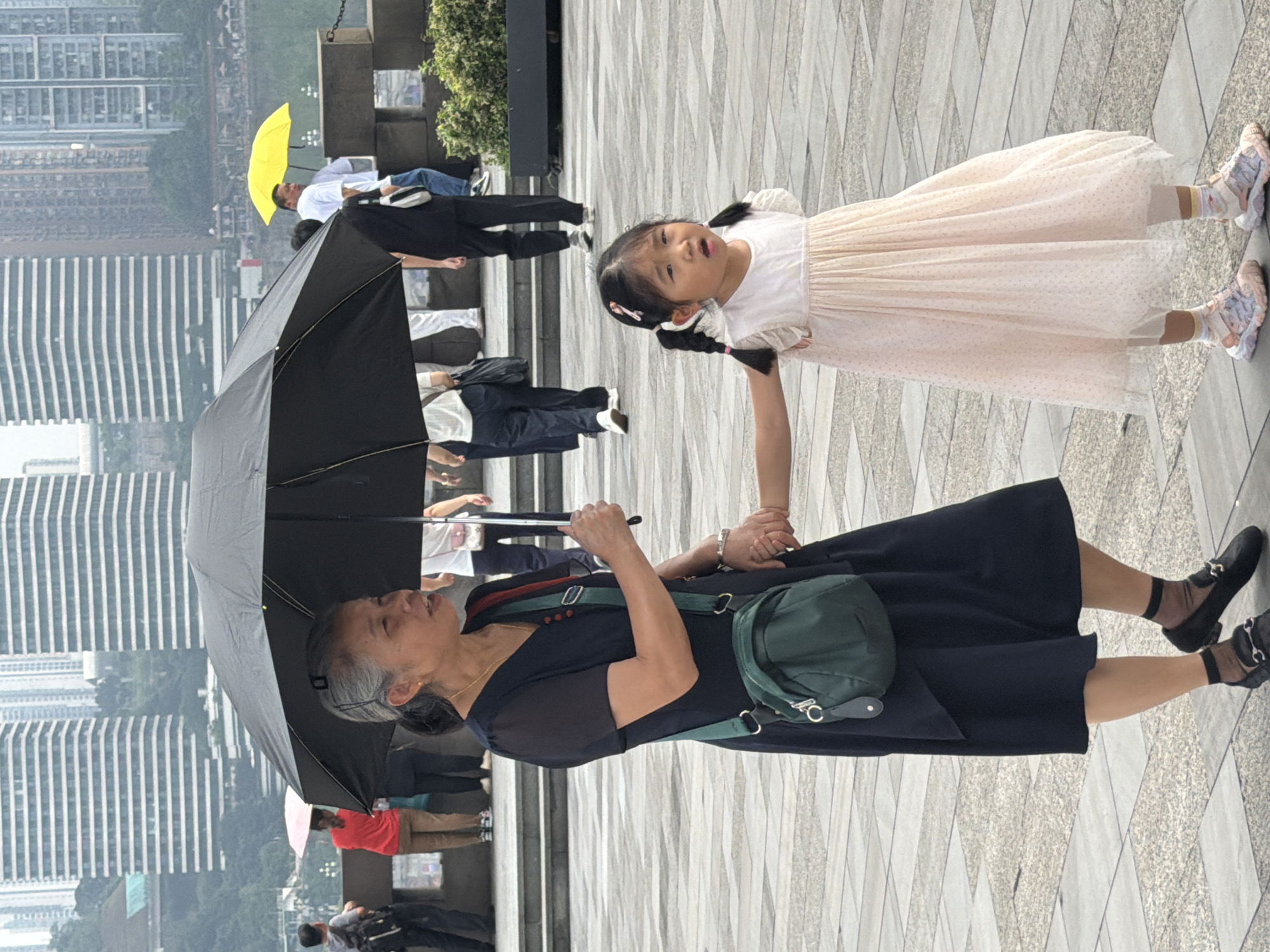 In Yuzhong District, China, a woman and a young girl are walking side-by-side under a large black umbrella. The woman, with gray hair pulled back and wearing a dark dress, holds the girl's hand. She is also carrying a dark green crossbody bag. The girl, wearing a pale pink dress with small patterns and white socks, looks towards the camera with an open mouth. In the background, several tall, modern buildings with horizontal striped facades dominate the cityscape. Other people are visible in the distance, some holding umbrellas, including a bright yellow one. The ground is paved with square tiles.