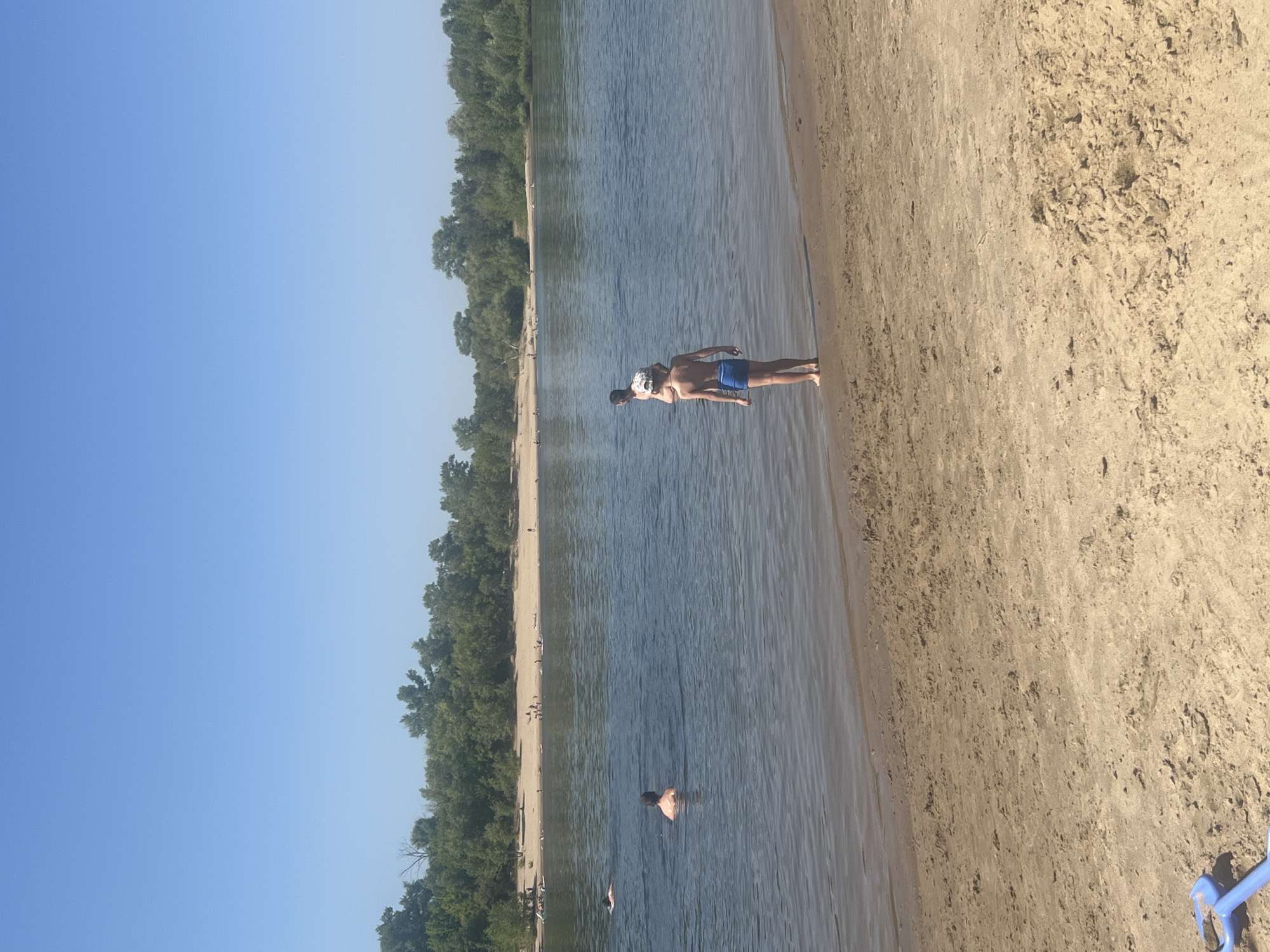The image depicts a sunny day at a sandy beach on a body of water, likely a river, with a dense line of trees bordering the far bank. The scene is oriented vertically, with the sky at the left and the sand at the right.
Several people are visible. In the middle ground of the water, two individuals appear to be floating or swimming, one on their back with a patterned head covering. Further out in the water, another person is partially submerged. Along the shoreline and in the shallow water, other figures can be seen, some appearing to be sunbathing on the sand. The beach itself is a light tan color, with some darker sand and debris scattered across it. The water is a calm blue with slight ripples. The trees on the far bank are lush and green, suggesting a warm season.
The overall atmosphere appears relaxed and recreational, characteristic of a summer day at the beach. The lighting suggests it is daytime, likely mid-morning or afternoon, with the sun casting a warm glow on the sand and water. There is no visible text in the image. The presence of a blue plastic toy shovel on the sand on the right side of the image adds a subtle detail of a family or children's outing.