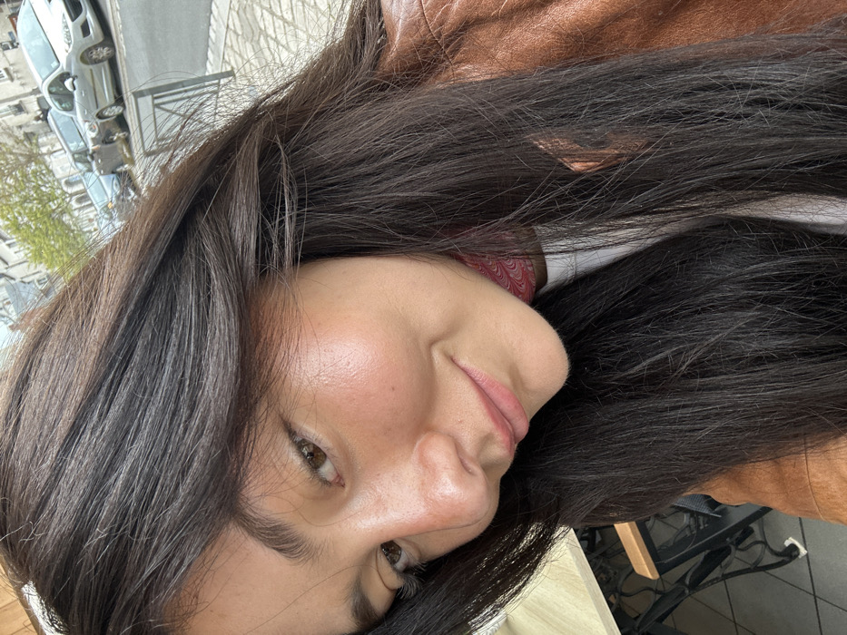 The image shows a close-up of a woman's face. The woman is looking down and to the right. She has long, dark hair and is wearing a brown leather jacket. She appears to be smiling slightly. The background of the image includes the lower part of the woman's white shirt and a blurry view of a city street. The woman's nose is the primary focus of the photo. The coordinates provided (latitude: 47.587, longitude: 1.337) indicate a location in Blois, France, but the image does not offer any visual clues to confirm this.