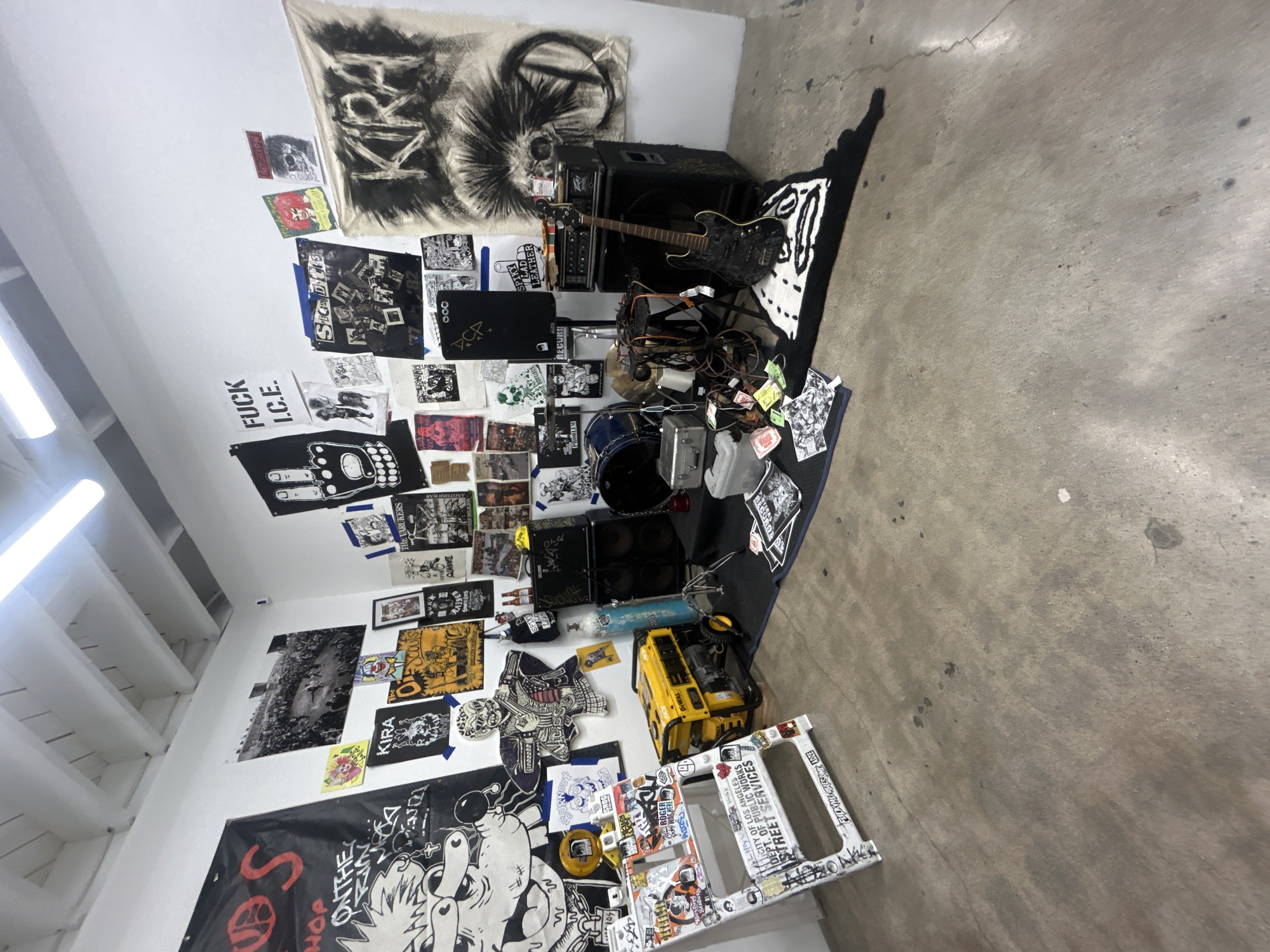 This image captures an indoor space, likely a practice room or DIY venue in Los Angeles, set up for a music performance or rehearsal. The room features white walls and a concrete floor, illuminated by overhead fluorescent lighting.
The central focus is a collection of musical instruments, including a black electric guitar resting against a Peavey amplifier stack, and a partial drum set consisting of a bass drum, cymbal, and other percussion elements, surrounded by tangled cables and microphones. A large yellow DeWalt power tool, possibly a generator, is positioned near the corner. A black and white patterned rug with graphic designs lies on the floor.
The walls are heavily adorned with numerous posters, fliers, patches, and fabric banners, creating a dense, anarchic aesthetic often associated with punk or hardcore music subcultures. Visible text on these decorations includes "FUCK I.C.E.", "KIRA", "SECTAL DILIS", "THE VARUKERS", "SPIKY SLAD LEATHER", "MYEKION", "ONTHE HOP", "SNOG", "ROGER WAS HERE", "ADZE", and "000". One prominent banner features a distressed, skull-like face. Another piece of wall decor is a large black and white image of a crowd.
On a white metal frame, stickers and graffiti are visible, including "CITY OF LOS ANGELES DEPT. OF PUBLIC WORKS STREET SERVICES", confirming the location. The overall atmosphere suggests an raw, underground, and politically charged music environment. No people are visible in the scene.
