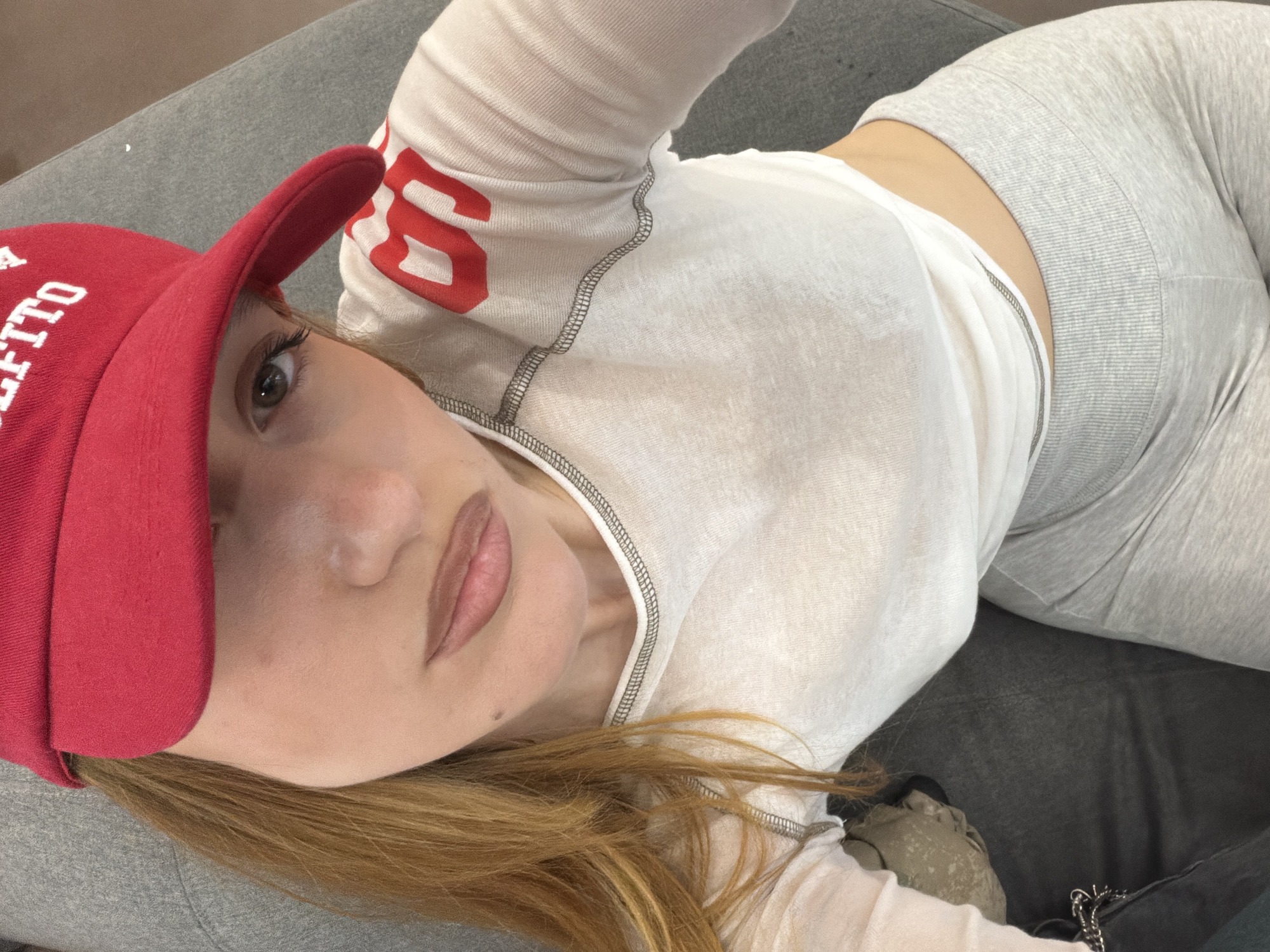 A young woman lies on a grey upholstered surface, possibly a sofa or large cushion. She wears a red baseball cap with white lettering, a long-sleeved white top with red lettering, and grey leggings. Her blonde hair is visible beside her face. The location is unknown and there is nothing to indicate that it is Sofia, Bulgaria.