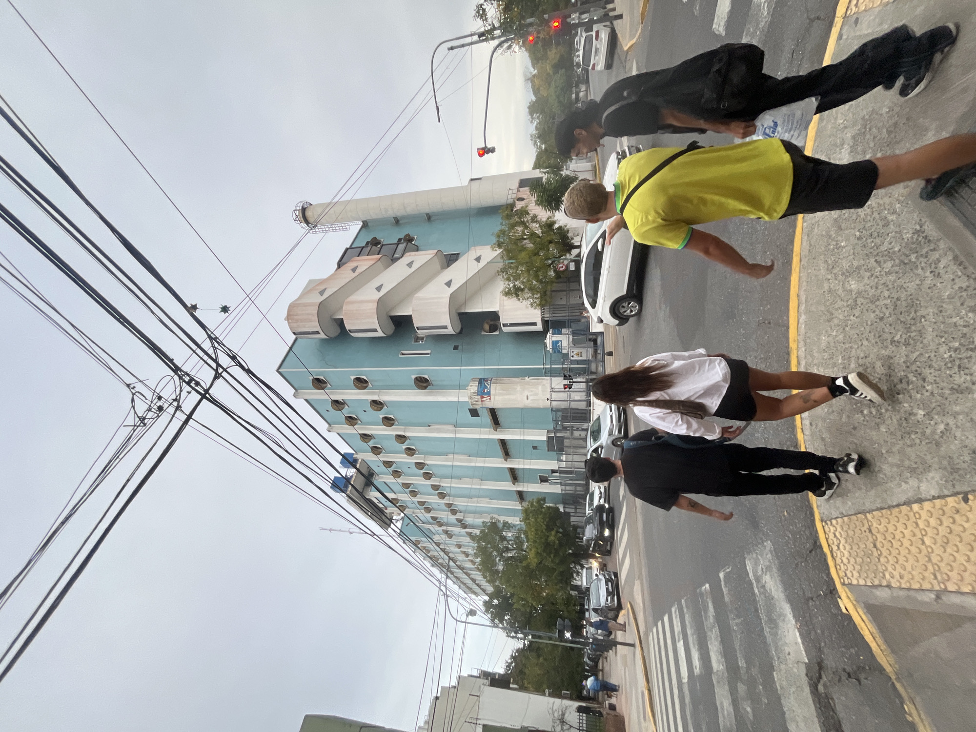 The image captures a street scene in Buenos Aires, Argentina. A group of four people is crossing a pedestrian crosswalk at an intersection. One person wears a bright yellow t-shirt and black shorts. Another wears a white shirt with black shorts. The other two individuals are dressed in black clothing. Cars are stopped at a traffic light, which is red. A tall, light-blue building with a prominent smokestack is in the background. The building features rounded windows and stepped architectural details. Utility lines are visible in the sky, and the weather appears overcast.