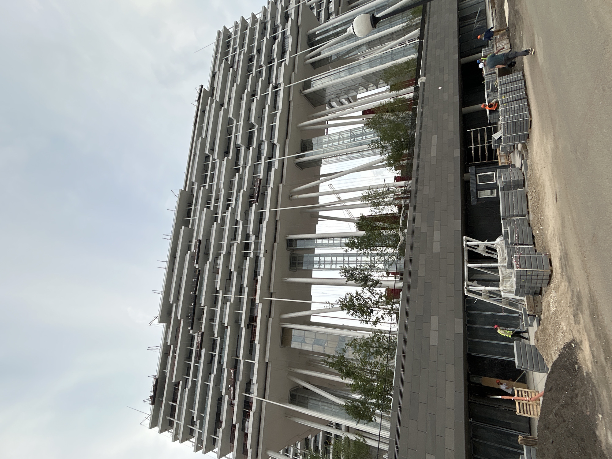 This is a low-angle, full outdoor shot of a large, multi-story building under construction in Moscow, Russia. The building features a distinctive architectural design with numerous staggered balconies and is supported by a forest of angled white pillars on the lower levels. The facade of the building is white and grey, with glass sections visible between the pillars. In the foreground, the scene depicts an active construction site. Several workers, dressed in construction attire and safety helmets, are present. They are engaged in various tasks, including moving materials, organizing construction supplies like pallets and stacks of paving stones, and operating equipment. The ground is a mixture of dirt, gravel, and paving stones. The sky is overcast. The overall impression is one of an ongoing construction project in an urban setting.