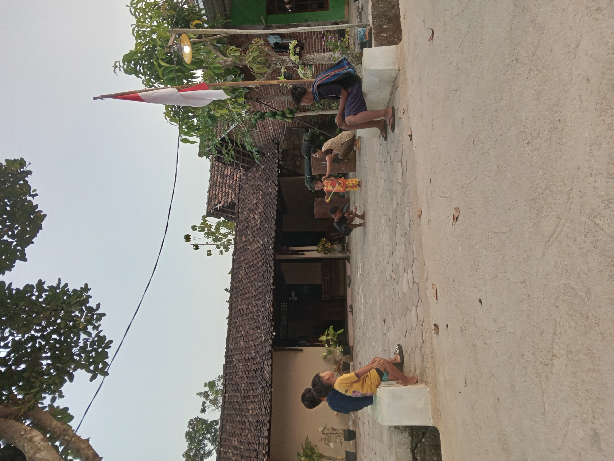 The image depicts a scene in the city of Kedunggudel, Indonesia, where several children and one adult are engaged in outdoor activities. A red and white Indonesian flag is prominently displayed on a flagpole. The environment features traditional Indonesian architecture with tiled roofs and brick walls. The ground is paved with hexagonal tiles in some areas and concrete in others, with a few scattered leaves indicating an outdoor setting.
In the foreground, on the left side of the image, two young boys are seated on a white ledge, facing away from the camera and towards a house. The boy on the right wears a yellow t-shirt and blue shorts, while the boy on the left wears a blue t-shirt.
In the middle ground, a group of three children and an adult are interacting. One child, wearing a yellow and orange outfit, appears to be playing a game, possibly with a string or a toy. Another child, wearing black clothing, is crouched on the ground. An adult, wearing a beige top and dark pants, is also in a crouched position, seemingly interacting with the children. To the right, a young person is seated on a similar white ledge as the boys on the left, facing the group in the middle. This individual wears a dark shirt and blue shorts and has a dark fabric draped over their shoulders.
The background reveals the residential structures of Kedunggudel, characterized by their rustic appearance. Trees, some bearing fruit, are visible around the houses, and a street lamp is mounted on a pole near the flag. The overall lighting suggests it is late afternoon or early evening, with the sky appearing bright and overcast.