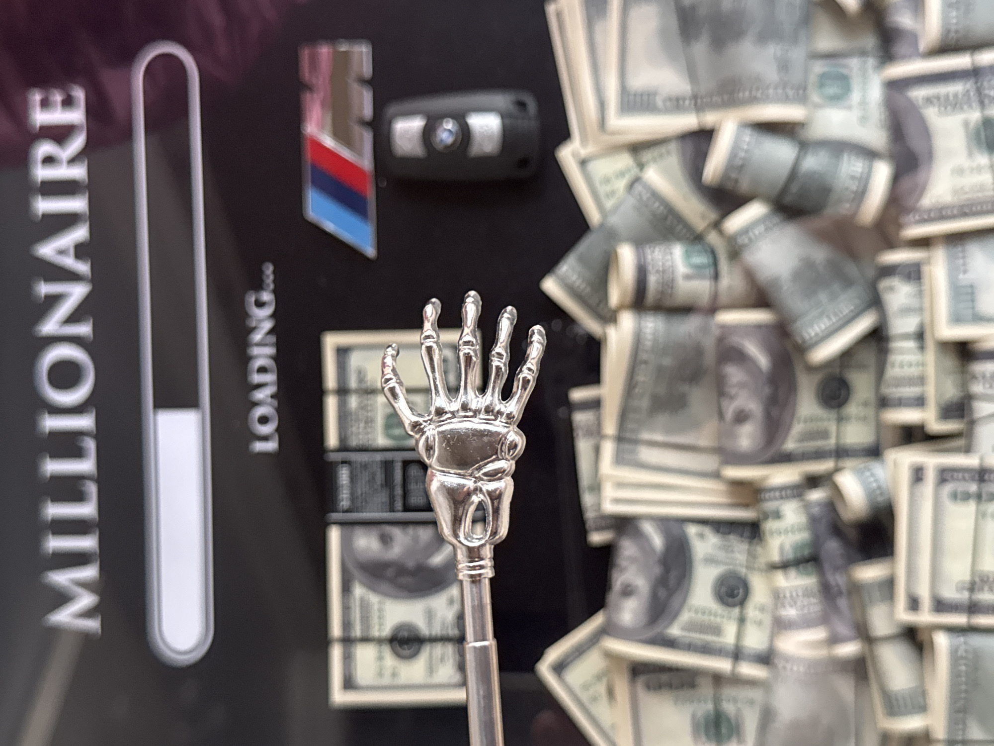 The image displays a black surface with the word "MILLIONAIRE" in large, metallic letters at the top, followed by a loading bar. Below the loading bar, the word "LOADING..." is visible. To the right, there is a BMW M logo and a car key fob. The scene is cluttered with stacks and rolls of US dollar bills, some of which are partially visible. A silver skeleton hand object is positioned horizontally across the middle of the frame, appearing to grasp some of the dollar bills. The background is dark and slightly blurred. There is no indication of the city of Yekaterinburg, Russia, or any specific environmental context beyond the objects themselves.