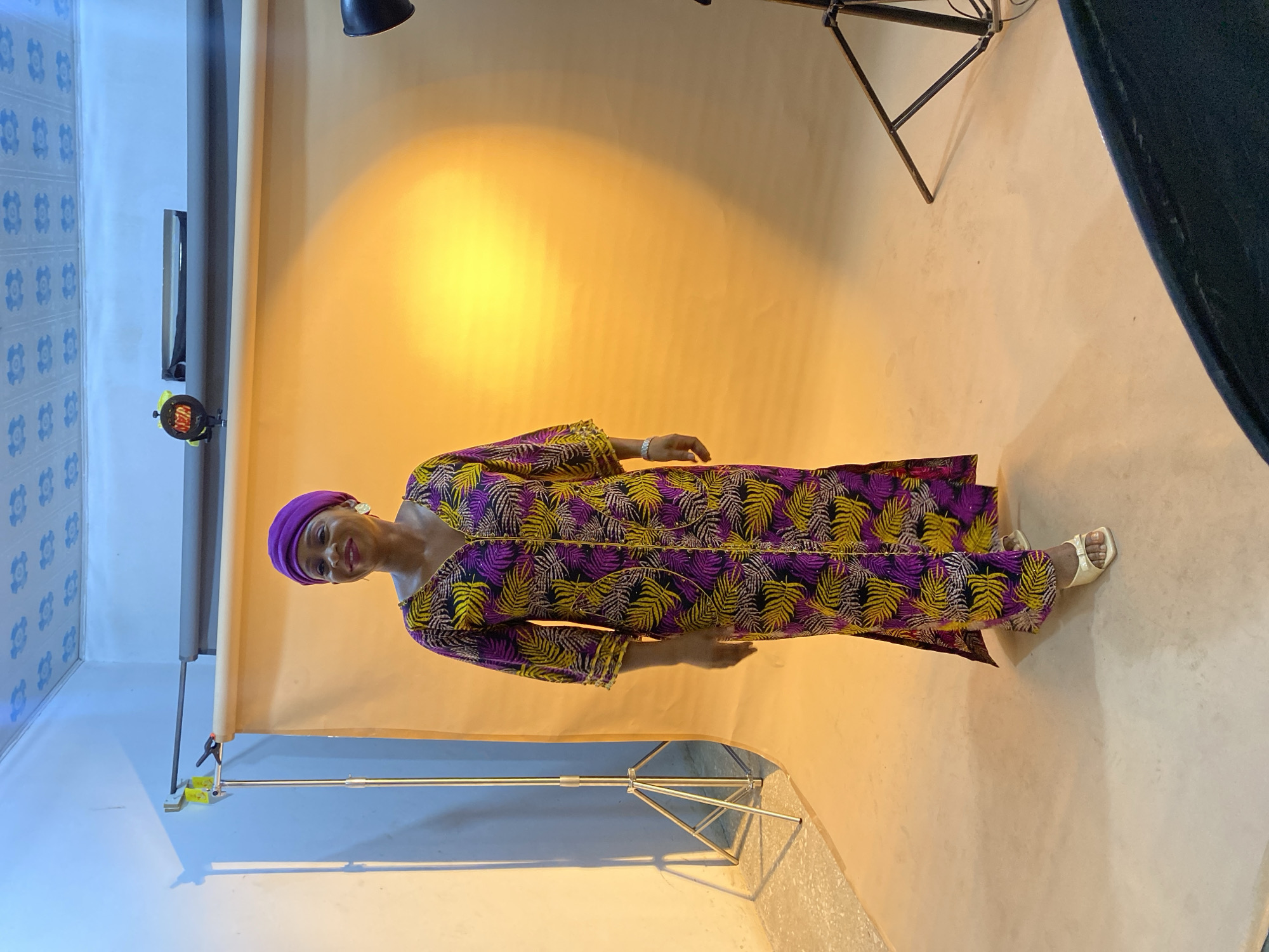 A woman stands in a photography studio. She is wearing a purple head wrap and a long dress with a purple, black, and yellow leaf pattern. She has light skin and is smiling. Her left hand is visible with a bracelet. She is wearing white sandals.
The background is a curved beige paper backdrop. A light stand with a black light is positioned to her upper left. Another light stand is to the right, partially visible, and a third is visible to the lower left. The walls of the room are light blue.
The studio is located in Gura Top, Nigeria.