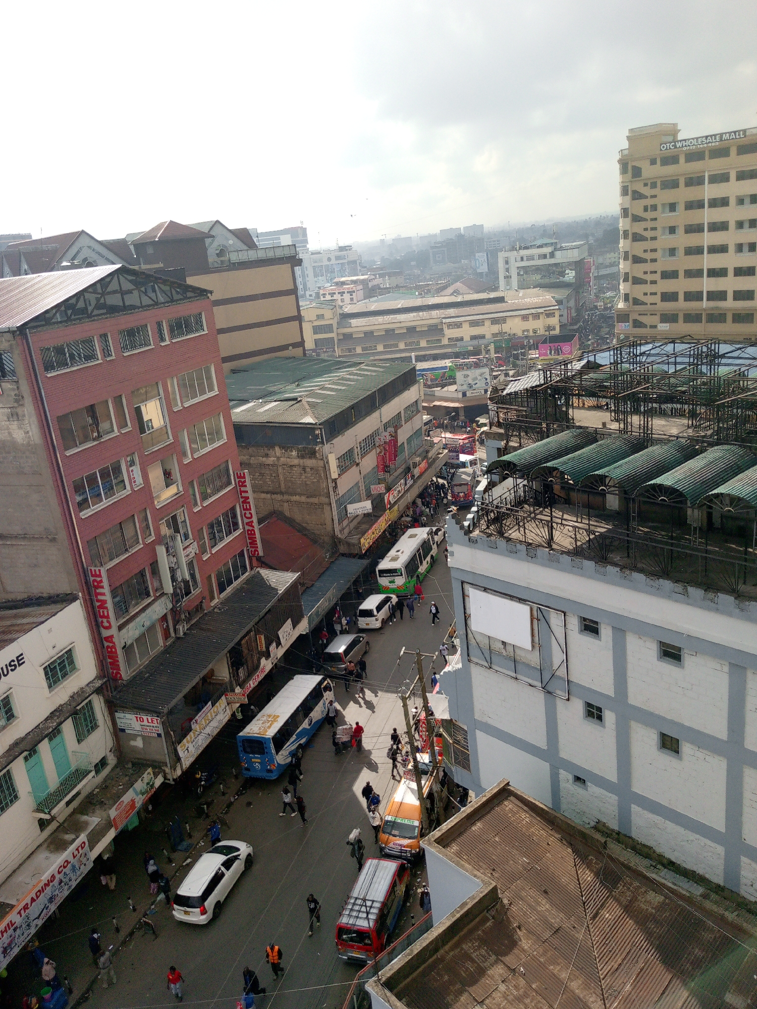 The image shows a busy street scene in Nairobi, Kenya, viewed from an elevated perspective. The street is lined with buildings, including the Simba Centre and OTC Wholesale Mall. Several vehicles are visible, including buses, cars, and vans, with people walking along the street and sidewalks. The environment suggests a bustling urban commercial district.