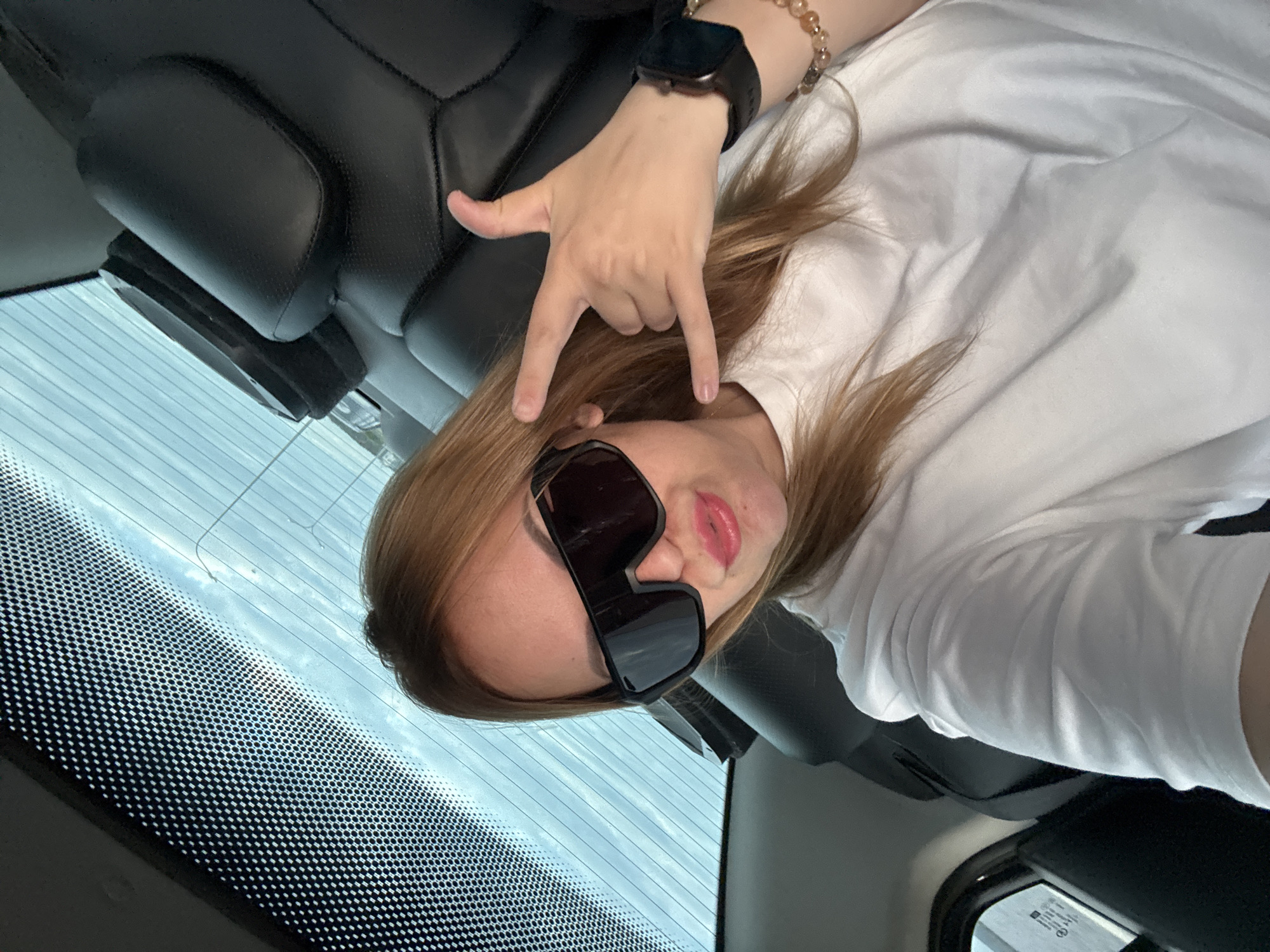 The image shows a woman in the back seat of a car. She is wearing large, dark sunglasses, a white t-shirt, and a bracelet. Her right hand is raised in a "rock on" gesture. The car's interior is visible, with black leather seats and a sunshade on the rear window. The scene appears to be in Chaikovsky, Russia.