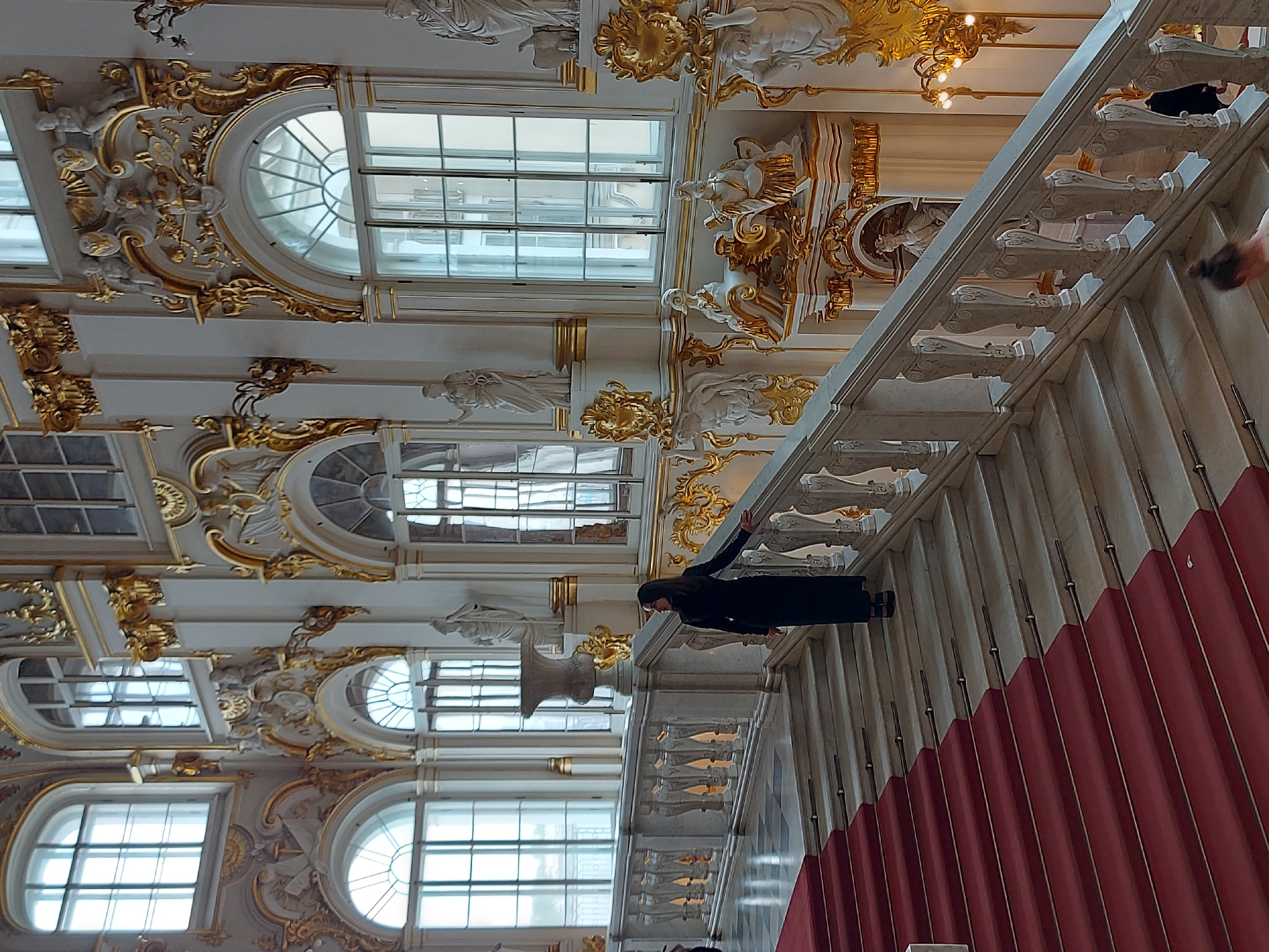 The scene, likely situated in Saint Petersburg, Russia, features an ornate interior with a series of large windows, decorative carvings, and a grand staircase. The windows are arched and rectangular, allowing ample natural light into the space. The walls and ceilings are adorned with intricate gold and white carvings and statues. A woman wearing a black outfit is seen walking along the staircase landing. A red velvet-like material lines the base of the staircase, adding a touch of opulence. The staircase features decorative banisters. The overall environment suggests a historic and elegant building, possibly a museum or palace.