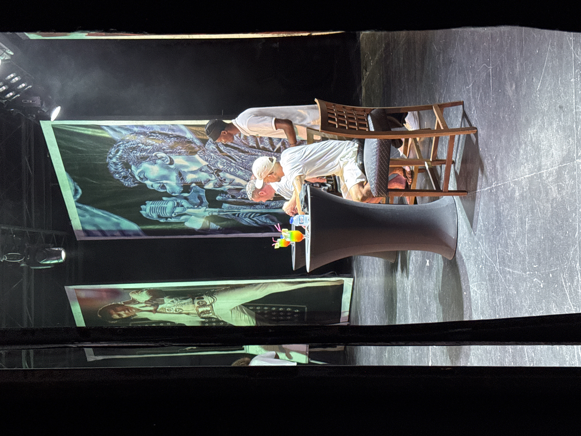 The image captures a stage set with three large portraits of musicians. On the left, a portrait of Elvis Presley is prominently displayed. To its right, a portrait of a Black male musician, possibly Bruno Mars, is visible. The stage floor appears to be a dark, possibly concrete or tiled surface, marked with various scuffs and lines.
In the foreground, three men are seated at high-top tables. The table on the left has two drinks with colorful garnishes. The men are dressed in casual, light-colored clothing, suggesting a relaxed atmosphere. One man in the center is wearing a white t-shirt and shorts, with a white baseball cap. To his right, another man in a white t-shirt and white shorts is seated. A third man, standing behind them, is wearing a white short-sleeved shirt and a black beanie.
The lighting appears to be stage lighting, casting shadows and highlighting the subjects. The overall impression is of a performance or event taking place on a stage, with a focus on musical icons and audience members. The location is not identifiable as Aksu, Turkey based on the visual information provided.