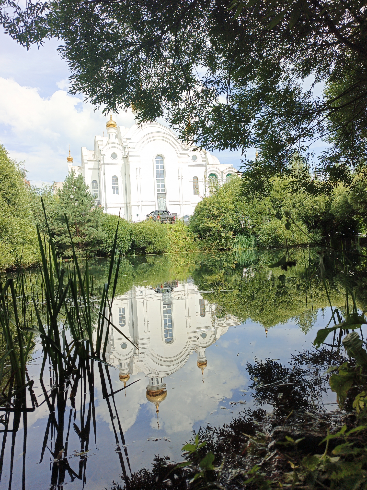 The image depicts a serene scene featuring a large white church with golden domes, reflected in a calm body of water. The church, identified as the Church of the Transfiguration in Zlatoust, Russia, is partially obscured by the lush green foliage of trees in the foreground. Reeds and aquatic plants grow along the edge of the water, adding to the natural beauty of the setting. The reflection in the water is clear, showing the church's architectural details and the sky above. The overall impression is one of tranquility and natural splendor.