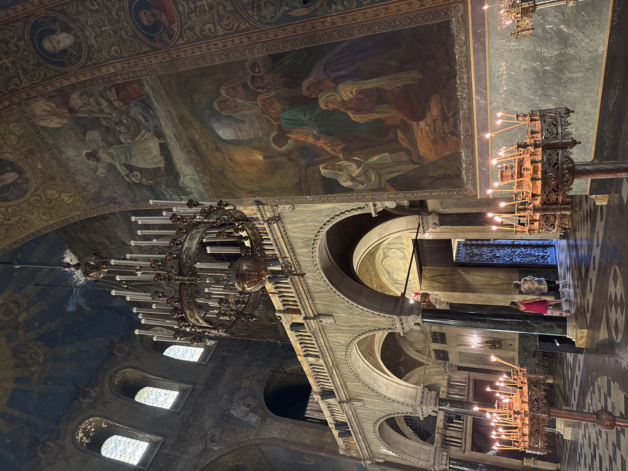 This image captures the interior of the Alexander Nevsky Cathedral in Sofia, Bulgaria. The view is a low-angle shot, looking towards the right side of the main hall.
The upper portion of the image reveals a vaulted ceiling adorned with dark blue frescoes depicting celestial figures and golden rays. Below the ceiling, multiple arched windows with geometric patterned glass allow natural light to enter the space. The walls are extensively decorated with large religious murals; a prominent fresco on the right depicts Jesus interacting with multiple figures, with another biblical scene located above it. Circular portraits within decorative frames are also visible on vertical bands along the walls.
A large, multi-tiered bronze chandelier with numerous electric candle lights hangs centrally from the ceiling, dominating the upper-mid section of the frame. Below this, an ornate marble balustrade, featuring numerous small columns, forms an upper gallery or balcony, supported by a series of grand arches.
On the ground level, large polished dark green marble columns with white bases and capitals rise to support the arches. The floor is composed of a distinctive black and white checkered pattern. In the foreground, two ornate metal candelabras stand, each holding multiple lit taper candles. To the right, another candelabra is partially visible.
Two adult females are positioned in the mid-ground on the right side. One woman, wearing a pink dress, walks away from the viewer towards a large, intricately designed dark metal door. Alongside her, another woman, appearing older and dressed in a patterned blouse and light trousers, also walks towards the door. A dark rectangular object, possibly a speaker, rests on the floor near a column.
