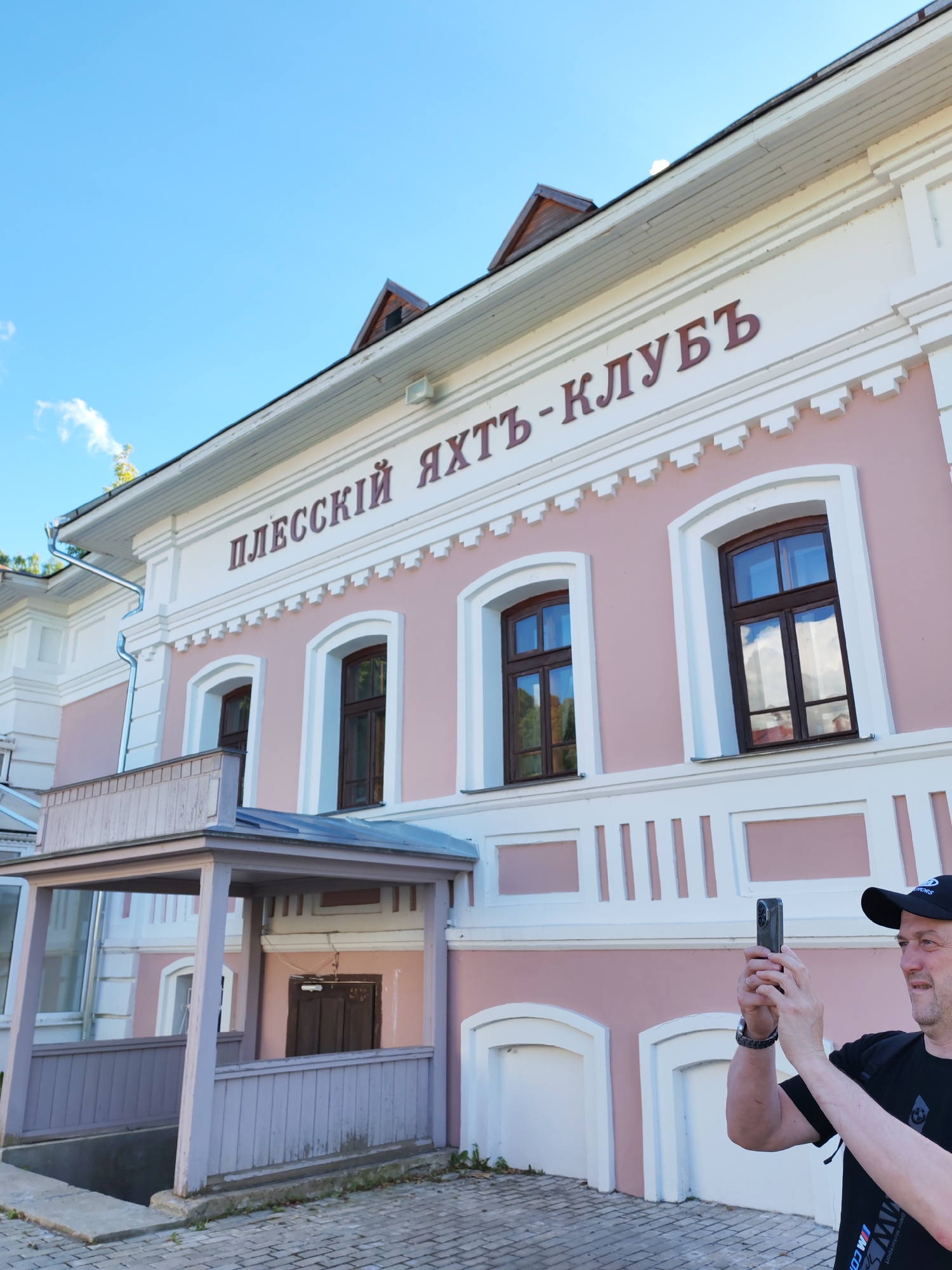 An outdoor, eye-level, medium shot captures a man in a black t-shirt and baseball cap holding up a smartphone to take a photograph in the city of Plyos, Russia. The man is on the right side of the frame and is looking to the right, slightly out of focus. The building behind him is a light pink structure with white decorative molding and dark brown windows. The words "ПЛЕССКИЙ ЯХТЪ - КЛУБЪ" are written in dark red lettering on the upper facade of the building. To the left of the man, a wooden porch with a railing leads down to an entrance. The sky above is clear and blue with a few white clouds. The ground in the foreground is paved with grey bricks.