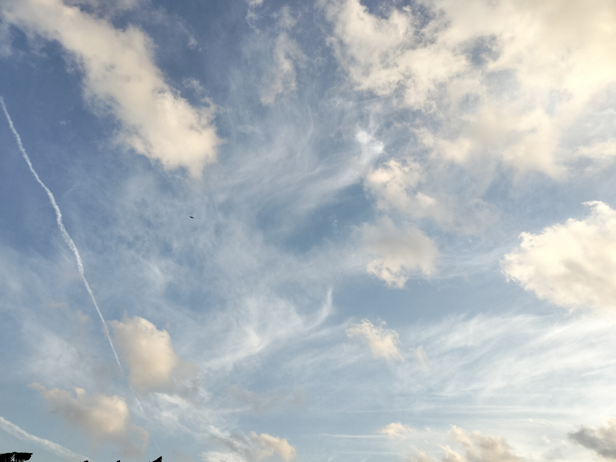 The image is a sky view from the city of Karang Indah, Indonesia. It features a blue sky with scattered clouds, some of which are white and puffy, while others are wispy and thin, creating a textured pattern across the sky. A distinct white contrail, likely from an aircraft, stretches diagonally across the upper left portion of the frame. In the center of the image, a small, dark object, possibly a bird, is visible against the sky. The overall impression is one of an open sky with light cloud cover, indicative of fair weather.