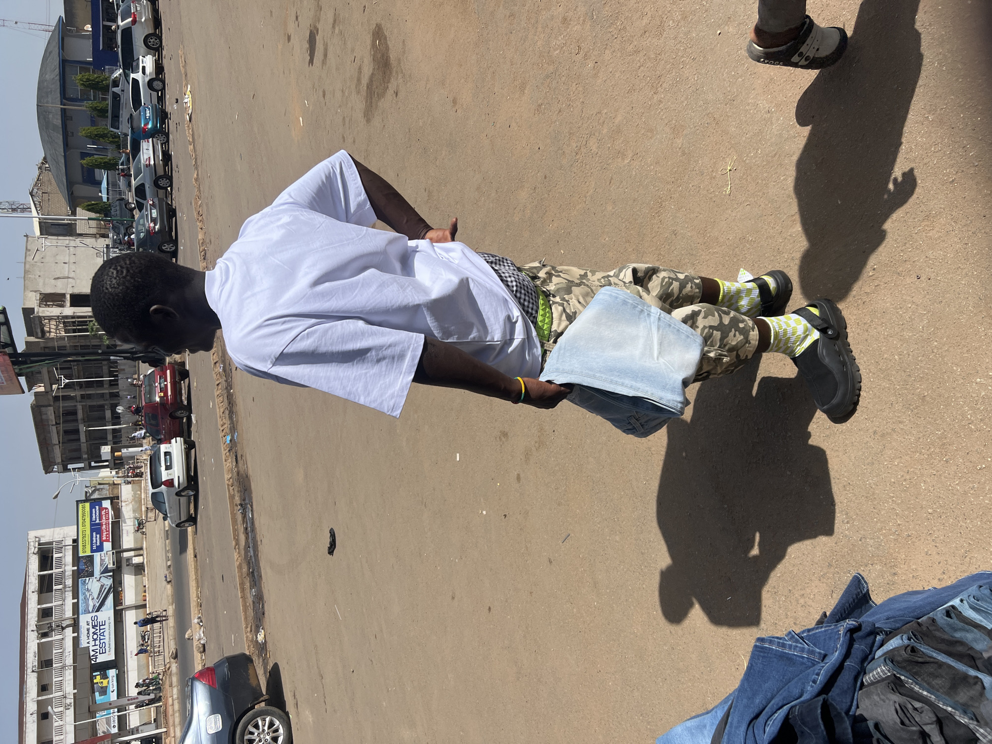 A medium outdoor shot depicts a man lying on his side on a brown concrete surface, possibly a sidewalk or road, in Jos, Nigeria. The man is wearing a white short-sleeved shirt, camouflage pants, and black Crocs with bright yellow and green patterned socks. He holds a stack of light-colored denim, possibly jeans. His left hand bears a yellow and green bracelet. To the lower right of the image is a pile of folded denim. The background includes a street with moving cars, billboards on a building, and a structure with a curved roof. A shadow is cast onto the ground near the man, and another pair of shoes is visible at the upper right corner.