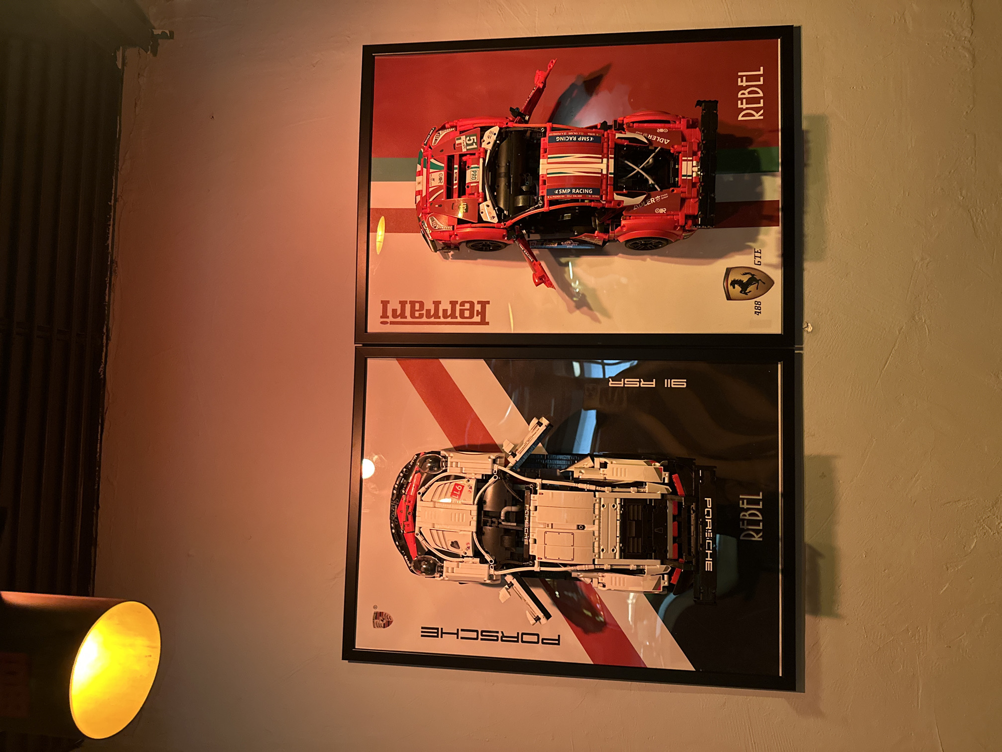 Two framed posters of LEGO car models hang vertically on a light-colored wall. The top poster depicts a red Ferrari racing car, seen from above, with its doors open. The name "Ferrari" is printed at the bottom. The poster also features racing stripes and the word "REBEL." The bottom poster features a white Porsche racing car, also seen from above, with its doors open. The word "PORSCHE" is printed at the bottom of the poster, along with "SII RSR" and the word "REBEL." A lamp is partially visible in the lower-left corner of the image, casting a warm glow. The location is likely indoors, given the presence of the framed posters and the lamp. There are no people visible in the image, and the location cannot be confirmed as Milan, Italy without additional information.