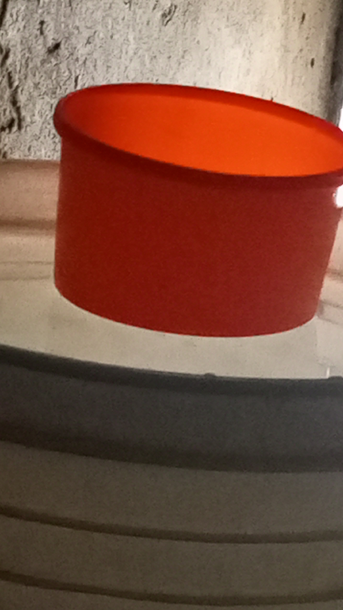 A red, cylindrical plastic container sits on top of a large, cylindrical, ribbed water tank. The water tank is gray near the top and transitions to a lighter tan color lower down. The background is a textured, light-colored wall. The location is Sabon Tasha, Nigeria.