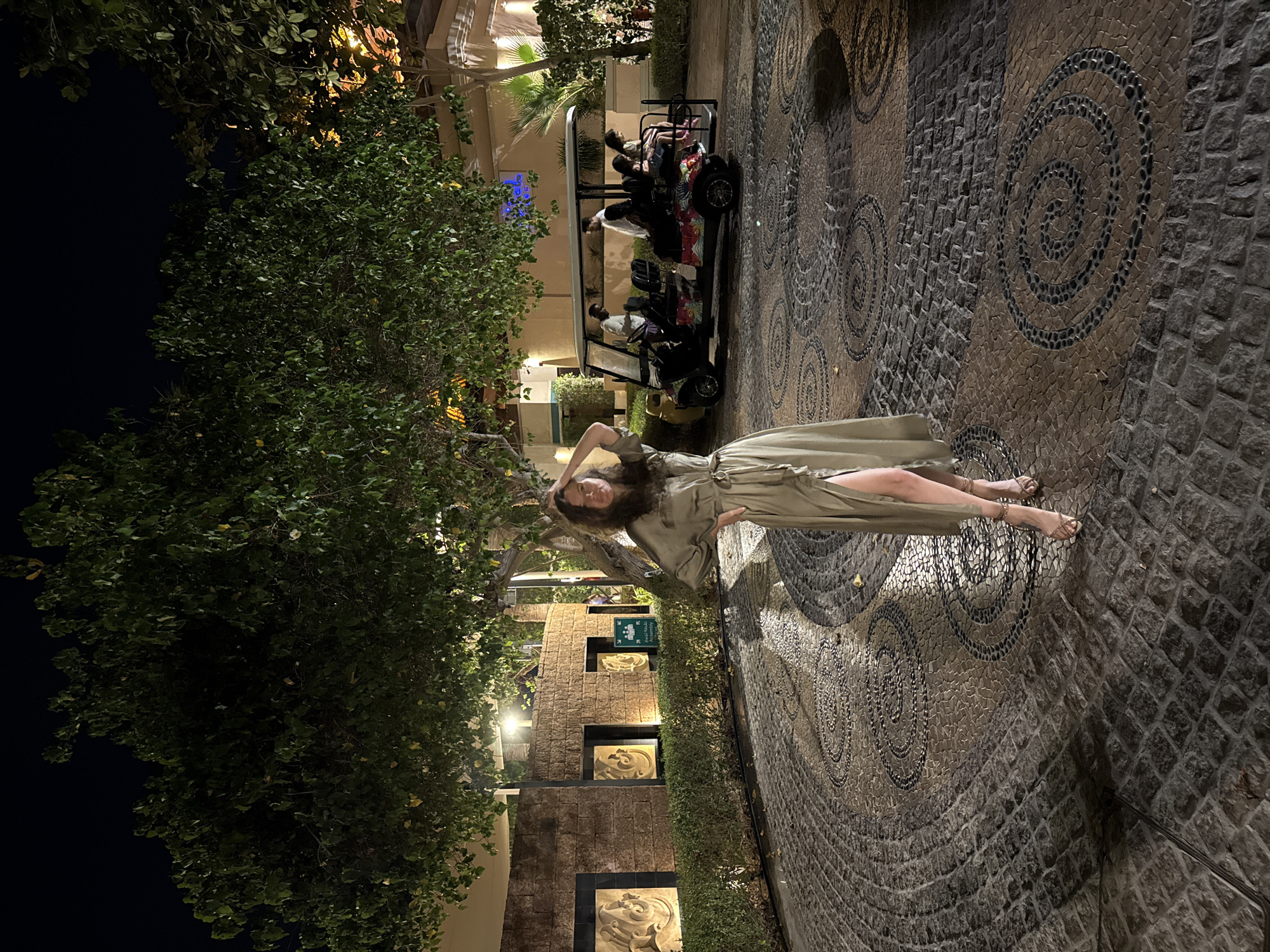 The image depicts a nighttime scene at what appears to be the entrance or a designated drop-off/pick-up area of a luxury resort or hotel in Dubai, United Arab Emirates.
In the foreground, a woman stands centrally, facing the viewer with a slight smile. She has long, dark, wavy hair and is wearing a long-sleeved, olive green midi-dress with a tie waist and a high slit on her left side. She is wearing light-colored, strapped high-heeled sandals. Her right hand is raised to her hair, while her left hand rests on her hip. The ground is paved with an intricate pattern: a central area features large circular mosaics created from dark and light-colored pebbles, set within darker grey cobblestone segments. The outermost perimeter is paved with uniform, rectangular, dark grey cobblestones.
To the left of the woman, a curved, textured stone wall structure is visible, made of light brown blocks. This wall incorporates several recessed square panels containing sculpted, swirling organic patterns. A green sign with white text and pictograms, indicating "نقطة تجمع / Assembly Point" with arrows pointing right and down, is affixed to this wall. Lush green hedges line the base of the wall, and dense trees with dark green foliage extend upwards, dominating the upper left and central background of the image.
In the midground, to the right of the woman, a multi-passenger golf cart (buggy) with a white roof and floral-patterned side panels is present. The golf cart contains five individuals: a male driver in a light-colored top, a male front passenger in a white long-sleeved shirt, and three passengers seated in the rear, two of whom appear to be women/girls in light-colored clothing. Buildings with warm illumination are visible in the background, including one with a reddish-brown tiled roof on the far right. The scene is illuminated by multiple artificial light sources, casting shadows and creating a warm, ambient glow across the paved area and architectural features. The sky above the tree canopy is dark, indicating night.
